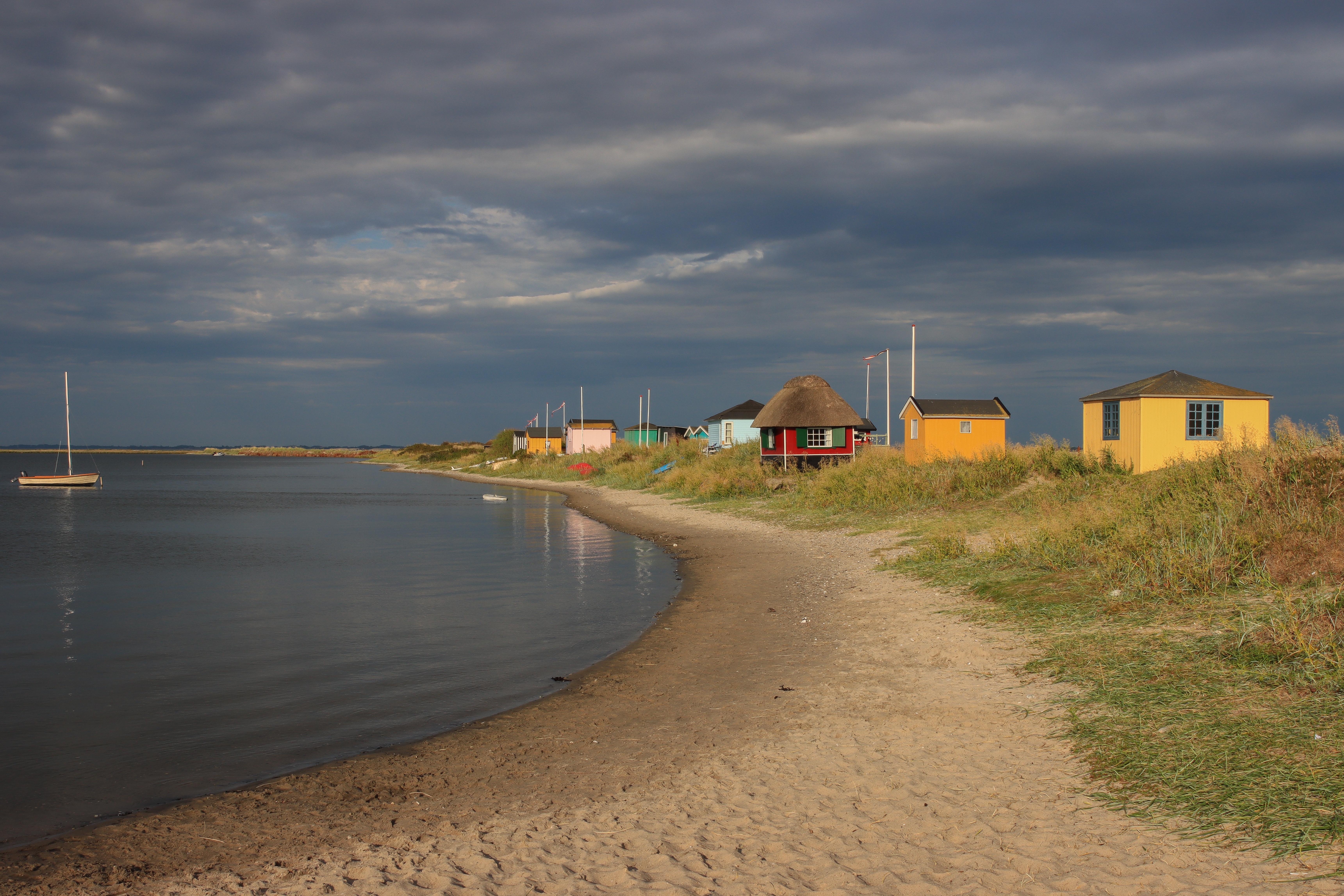 Idylliske farverige strandhuse ved Eriks Hale i Marstal på Ærø med roligt hav og en sejlbåd under en dramatisk overskyet himmel