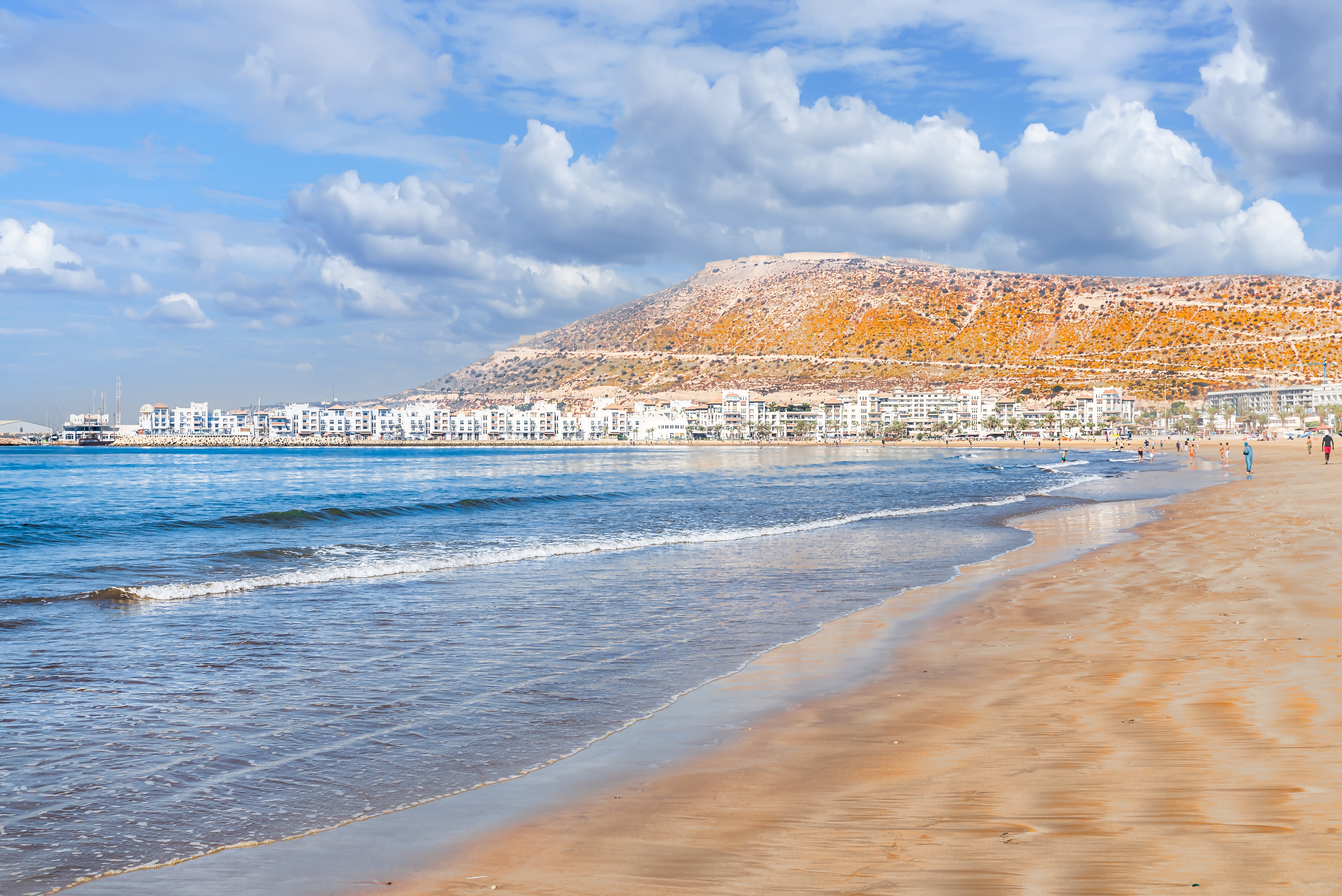 Idyllisk sandstrand i Agadir med blåt havvand, hvide feriehoteller og karakteristisk klippebakke under skyet himmel, Marokko
