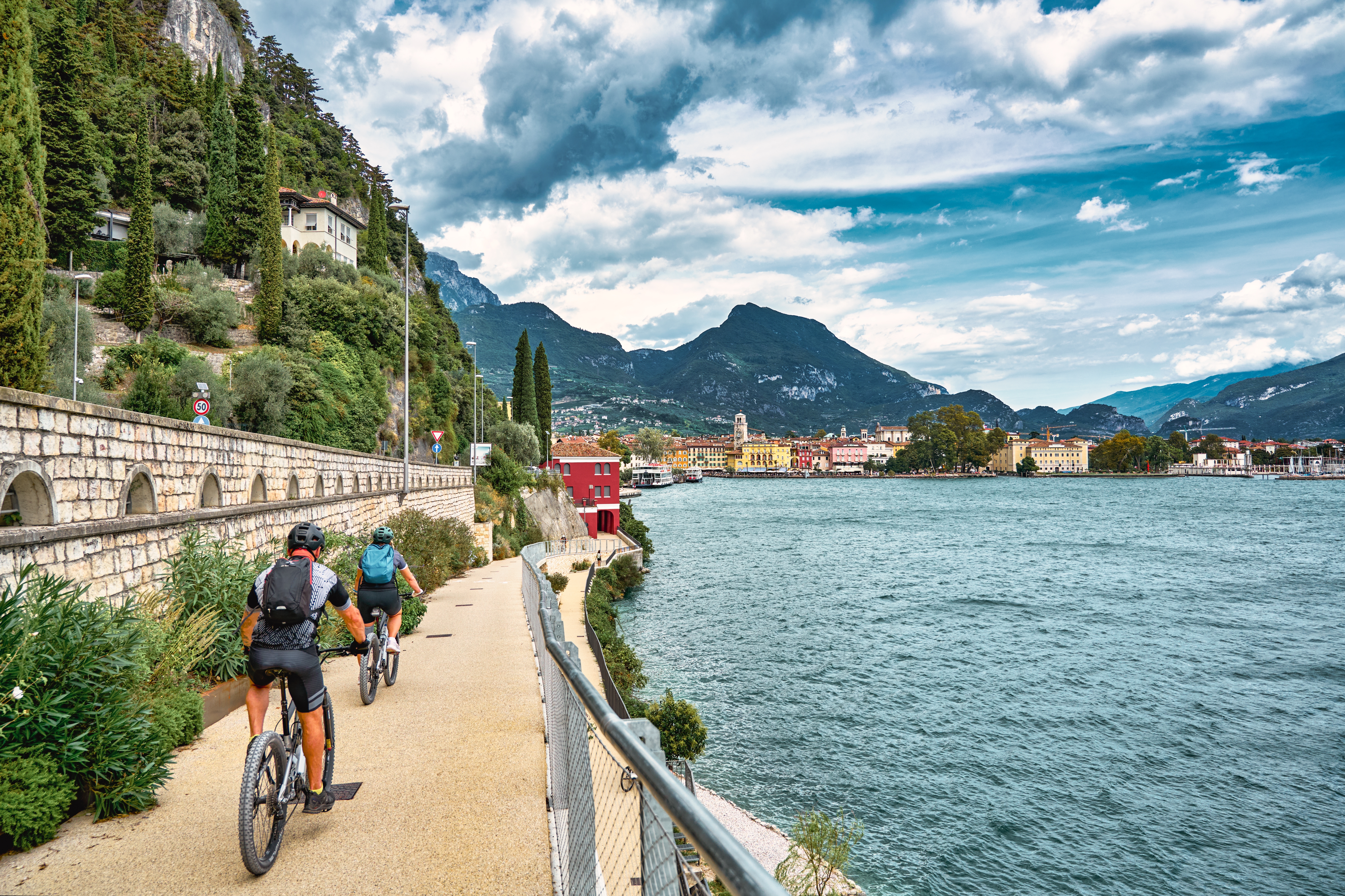 Cyklister på lakeside-sti ved Gardasøen med den pittoreske by Riva del Garda og dramatiske bjergtoppe i horisonten