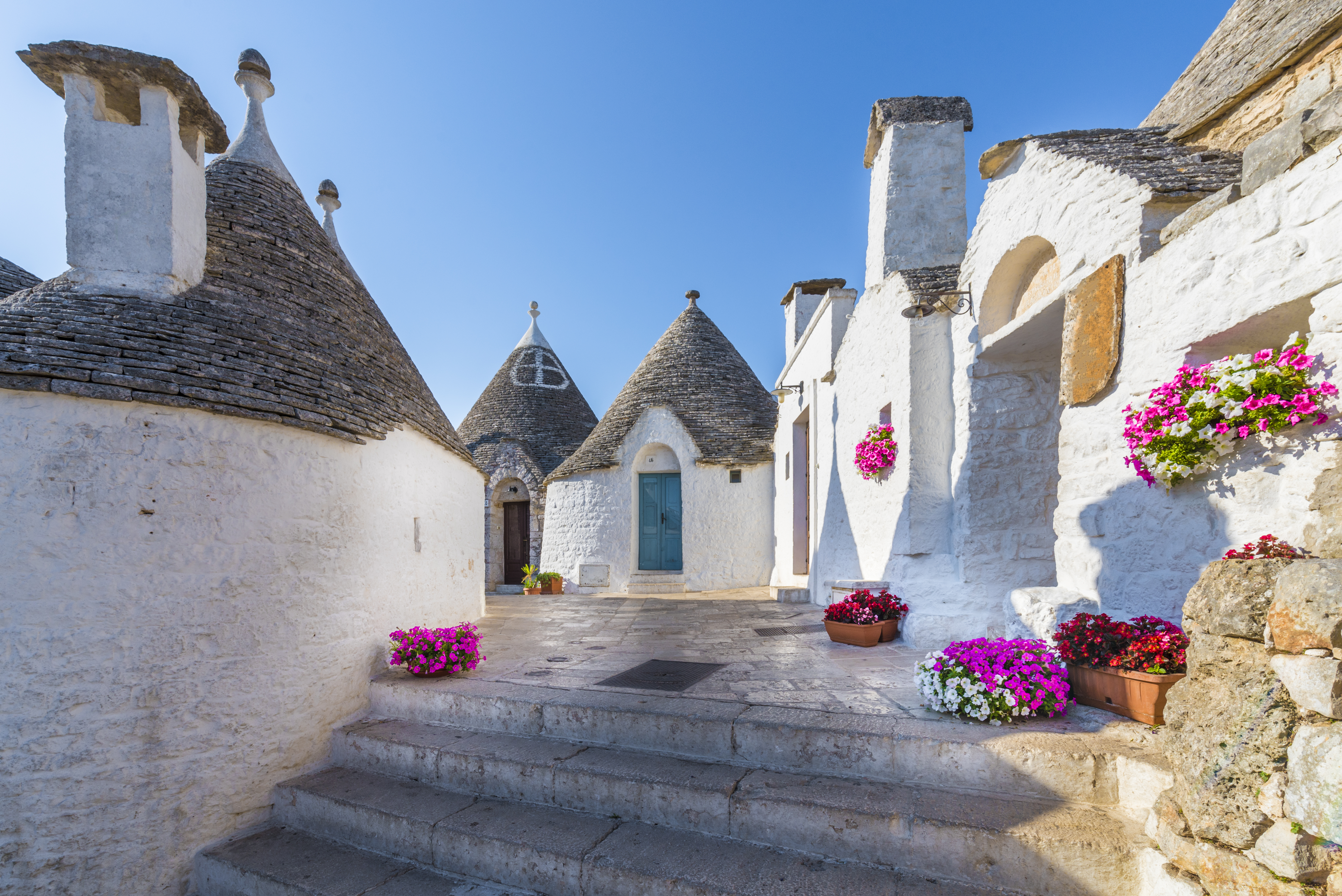 Et charmerende rejsebillede fra Alberobello med hvide trullo-huse og pink blomster under klar himmel.