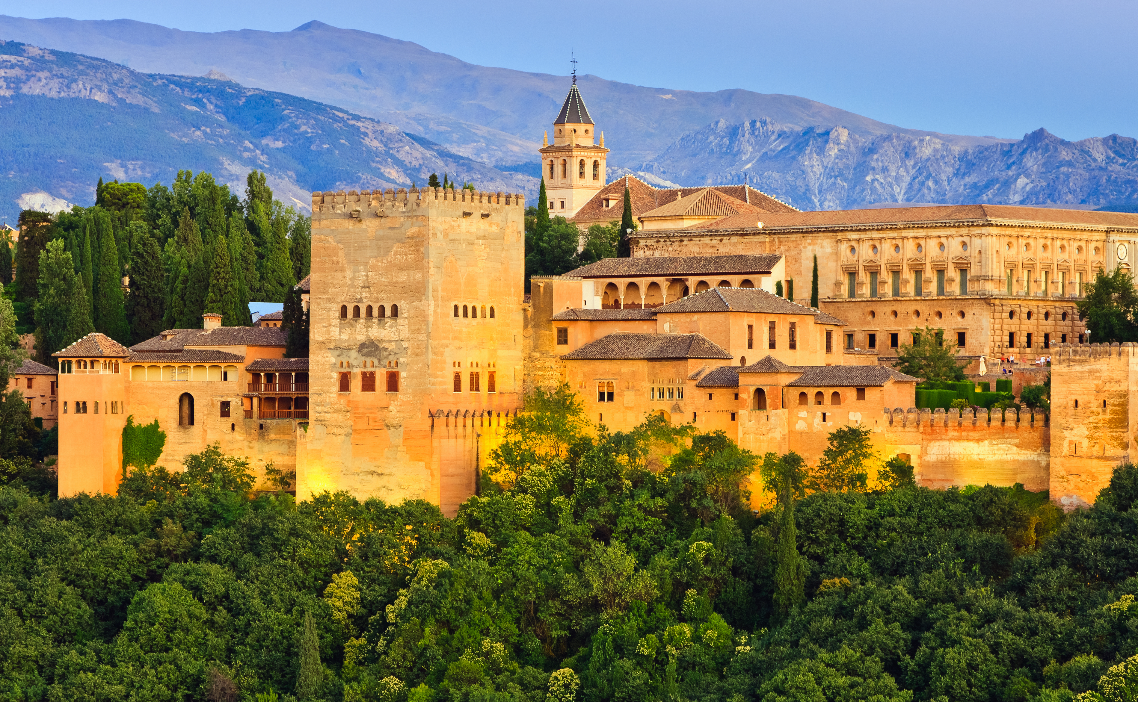 Den storslåede Alhambra-fæstning i Andalusien, Spanien, med dens karakteristiske gyldne mure oplyst i aftenlyset, omgivet af frodig vegetation og med Sierra Nevada-bjergkæden i baggrunden