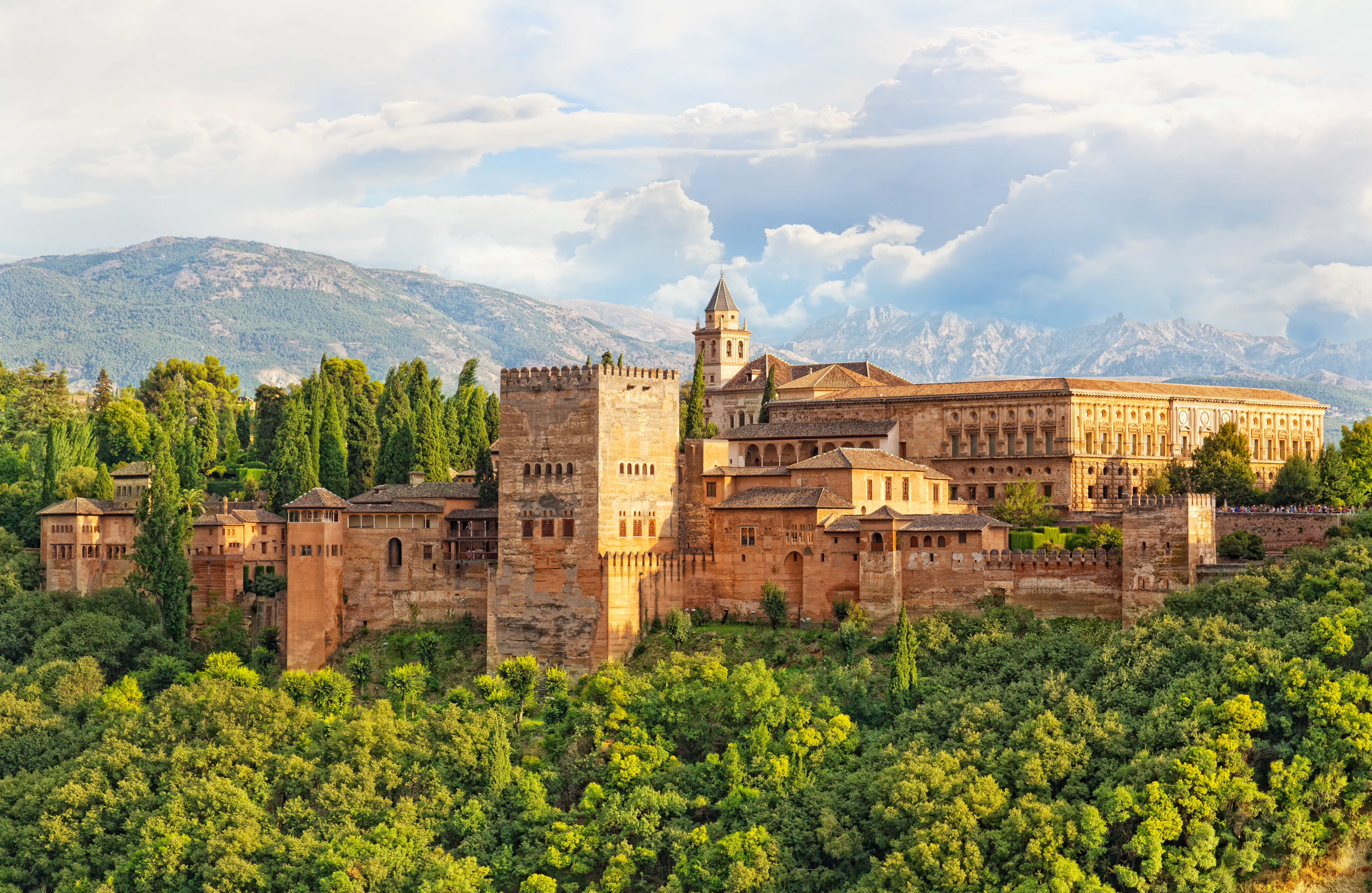 Den ikoniske Alhambra-fæstning i Granada med maurisk arkitektur og gyldne mure, omgivet af grønne træer med Sierra Nevada bjergene i baggrunden