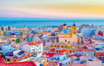 Farverig udsigt over Alicantes bymidte i skumringen med den smukke katedral med blå kuppel og det rolige Middelhav i horisonten på Costa Blanca, Spanien