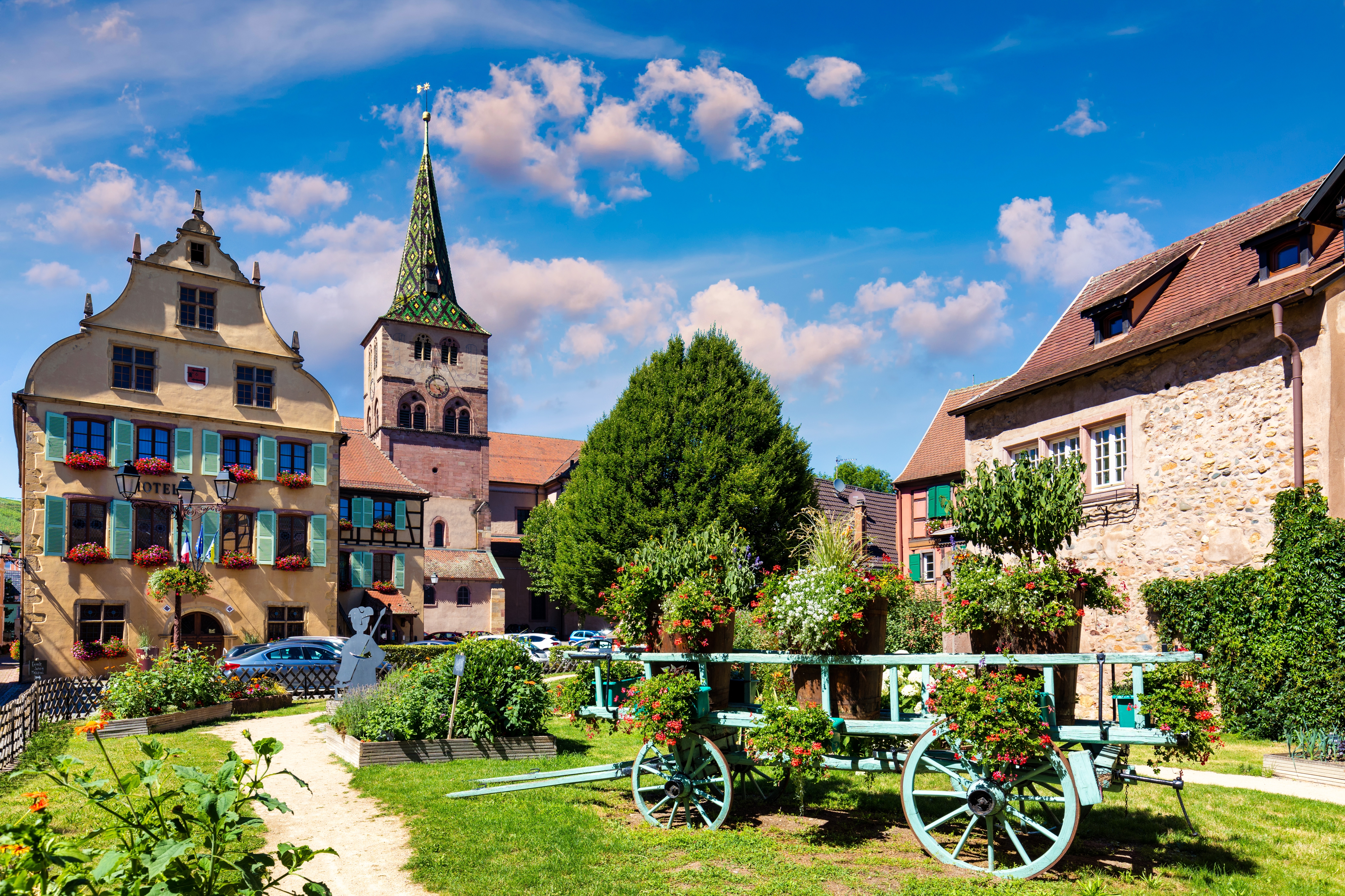 Stemningsfuld landsby i Turckheim, Alsace, med bindingsværksbygninger, blomsterdekoreret vogn og det karakteristiske grønne kirketårn i baggrunden