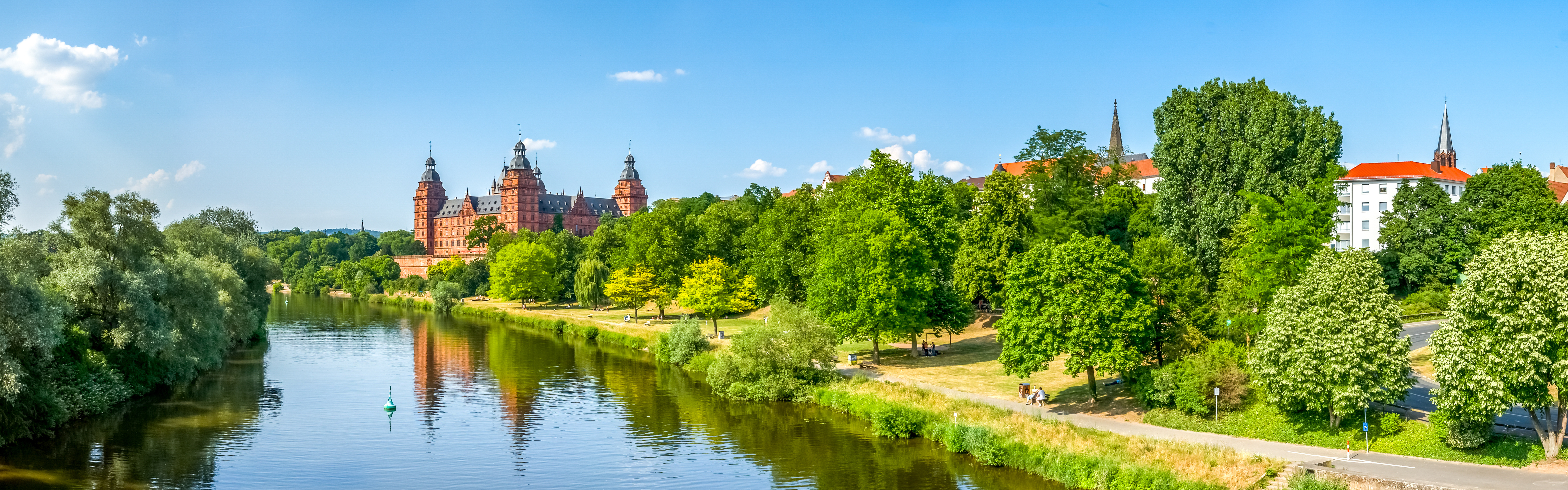 Schloss Johannisburg slottet i Aschaffenburg reflekteret i Main-floden, omgivet af frodige grønne træer og en klar blå sommerhimmel i Bayern, Tyskland