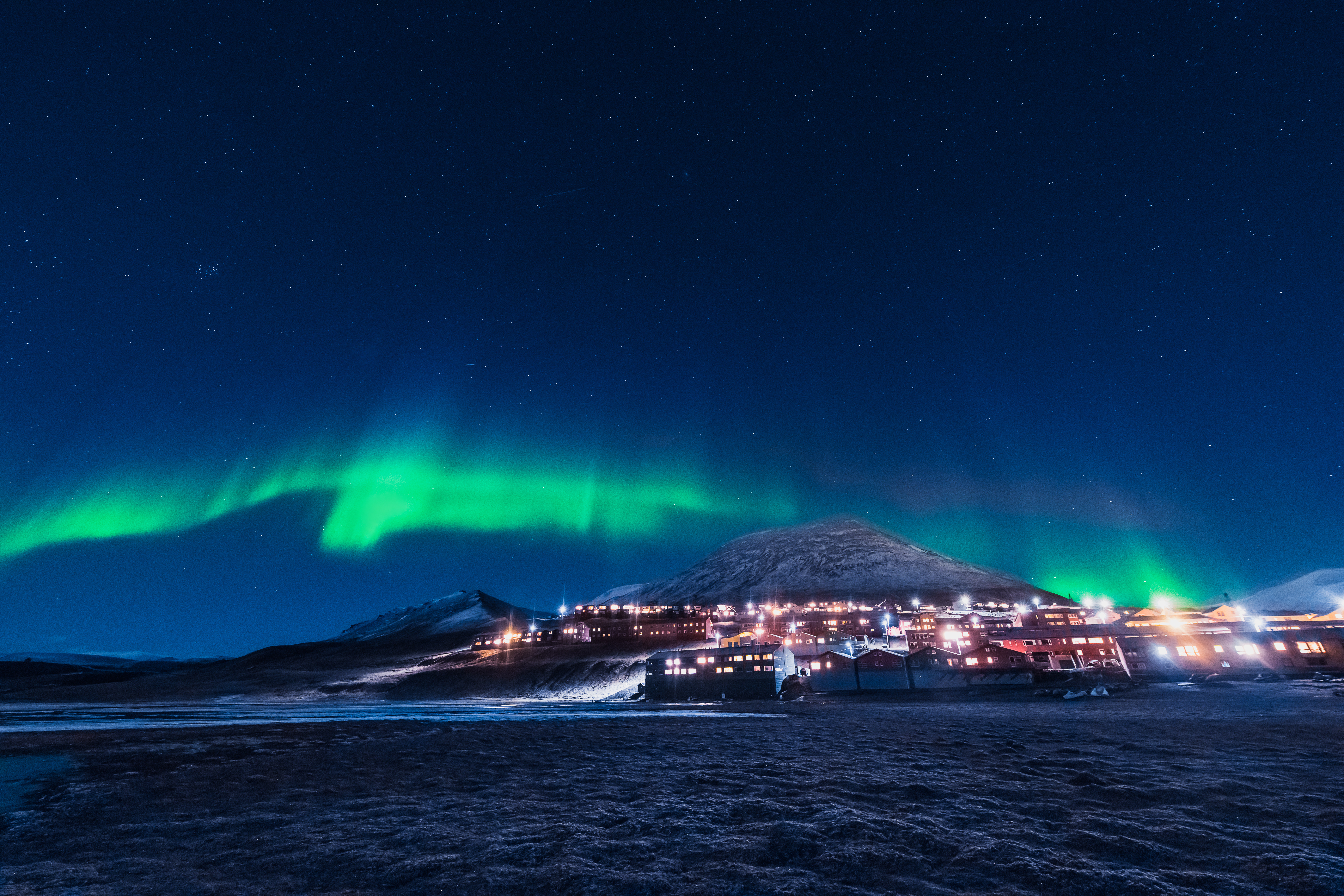Grønt nordlys aurora borealis over den oplyste by Longyearbyen på Svalbard i Norge med sneklædte bjerge og klar stjernehimmel om vinteren