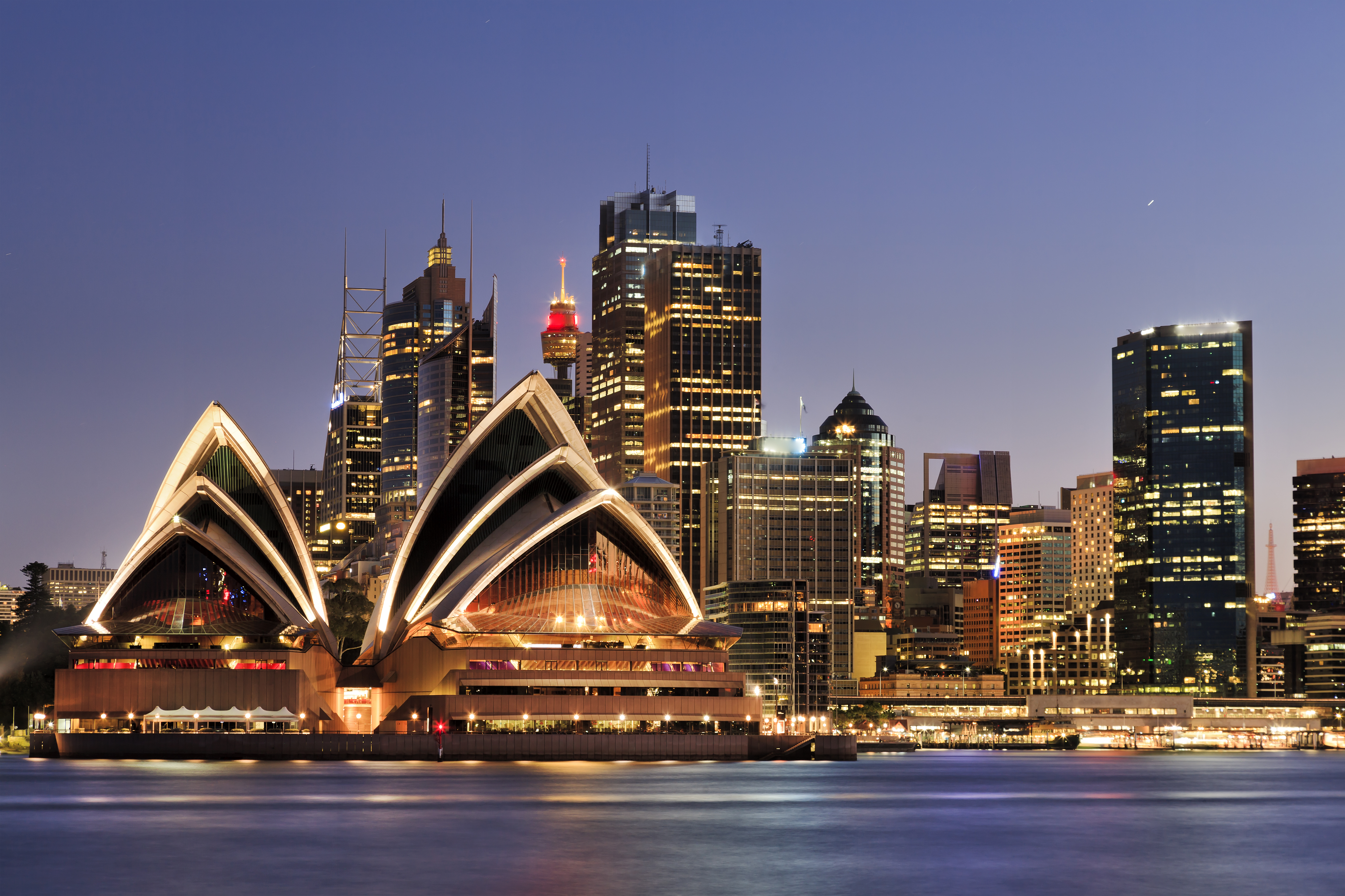 Det majestætiske Sydney Opera House med den oplyste skyline i baggrunden ved aftentid, spejlet i det rolige havnevand i Sydney, Australien