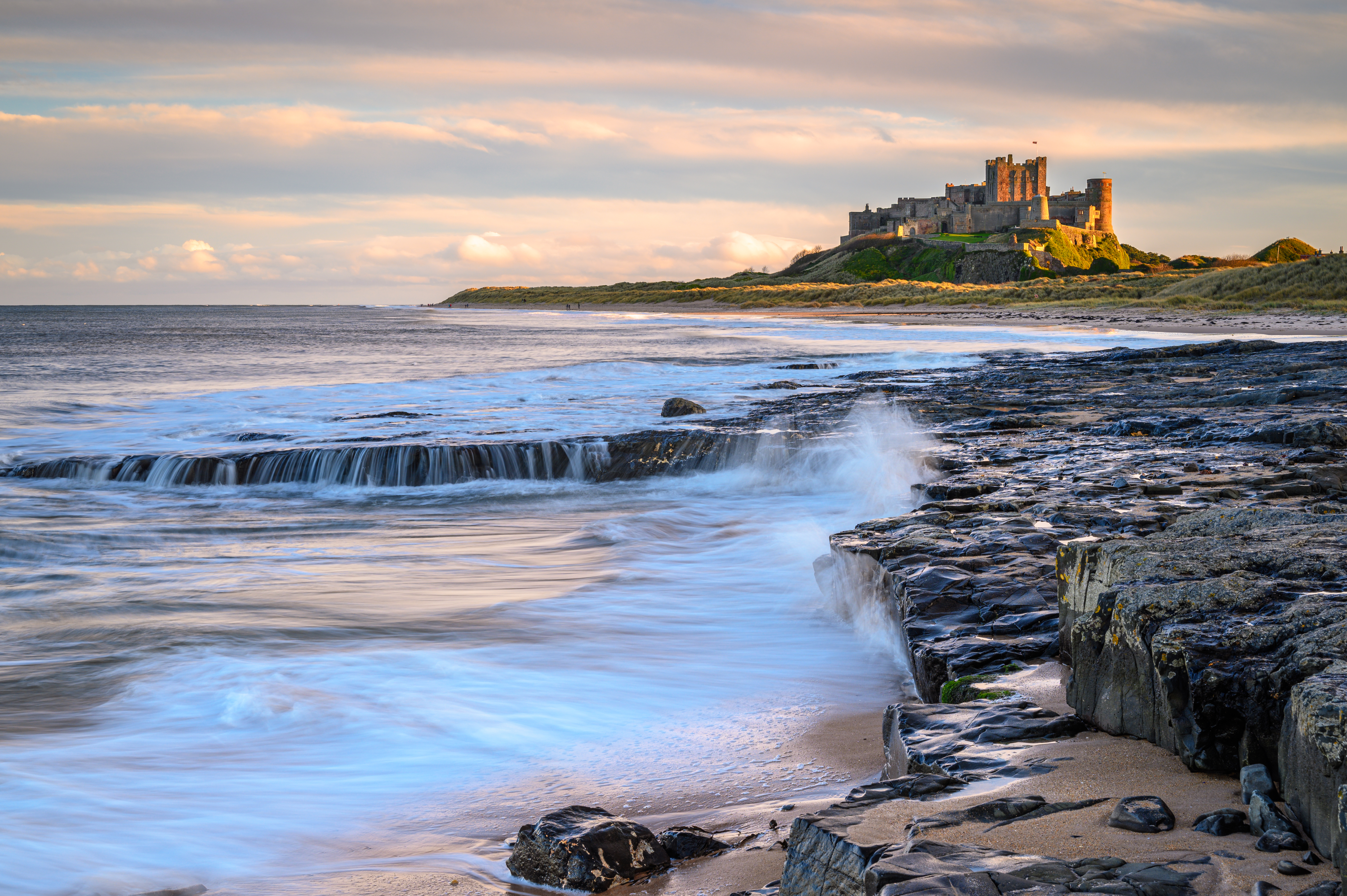 Udsigt til det majestætiske Bamburgh Castle fra stranden i Northumberland med bølger der bryder mod klipperne og dramatisk aftenhimmel