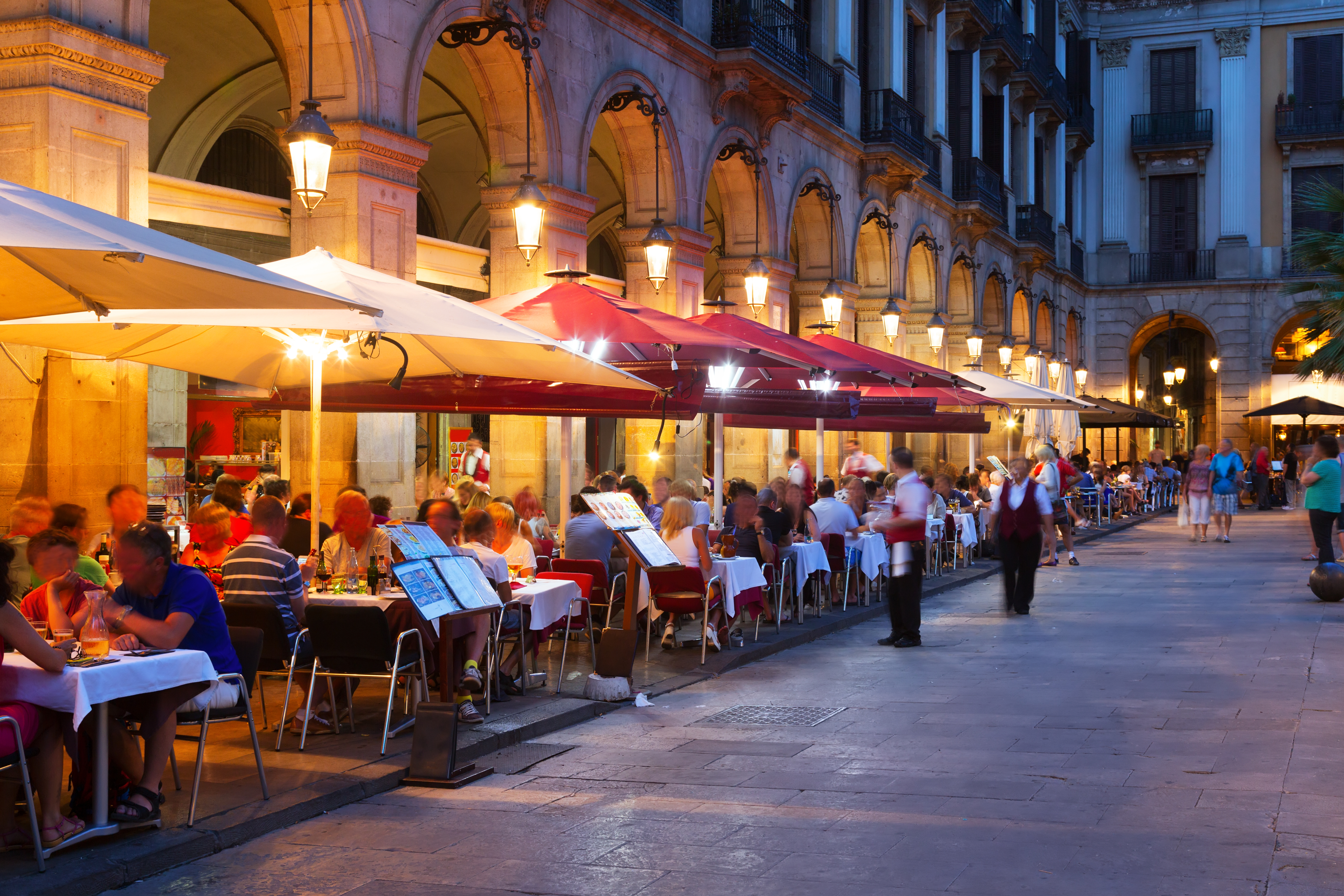 Livlig sommeraften på Den Kongelige Plads (Placa Reial) i Barcelona med oplyste restauranter, elegante arkader og den perfekte blanding af historisk arkitektur og moderne natteliv