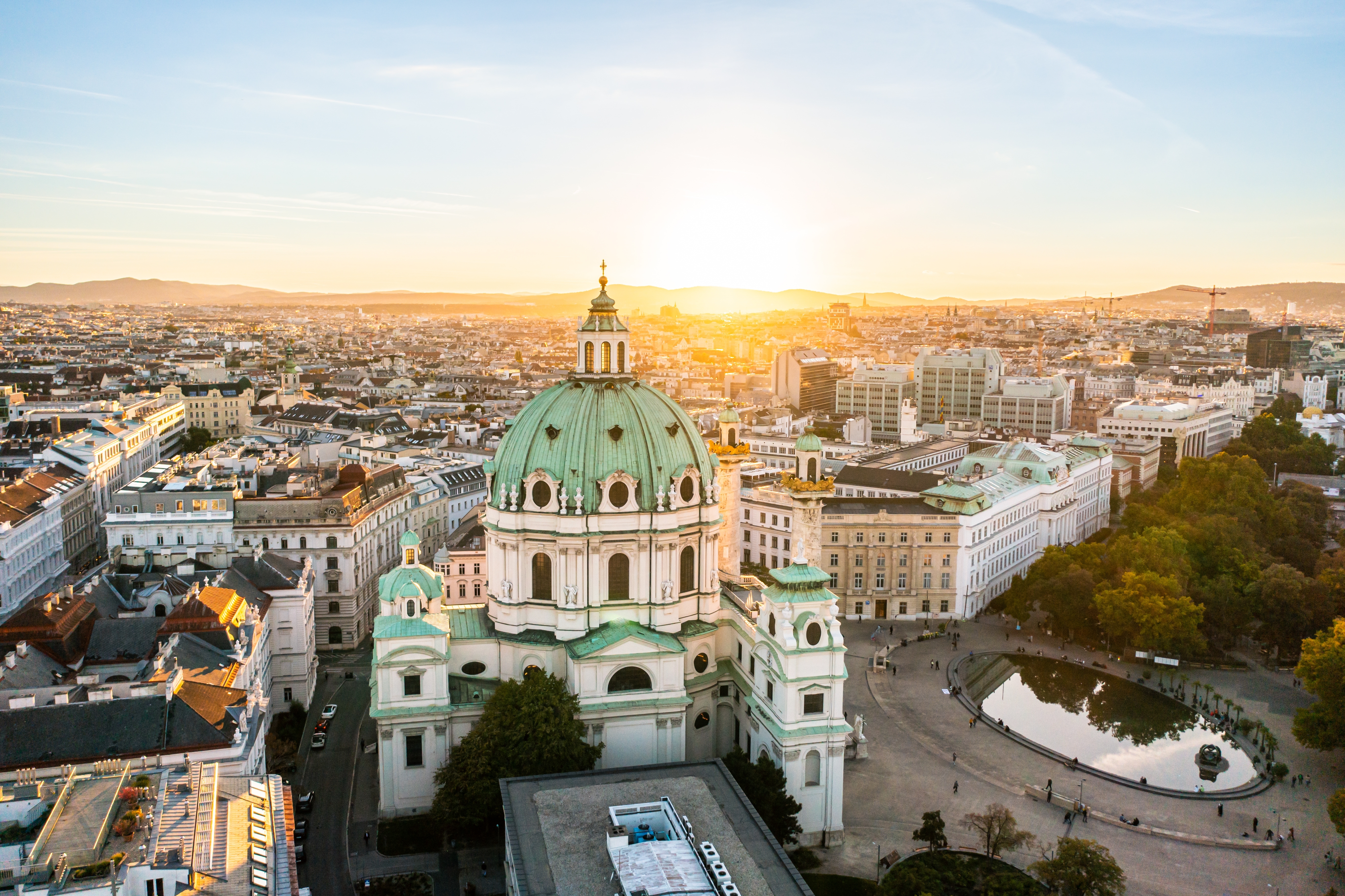 Barok St. Charles Kirke i Wien fotograferet fra luften ved solnedgang med byens landskab i baggrunden.