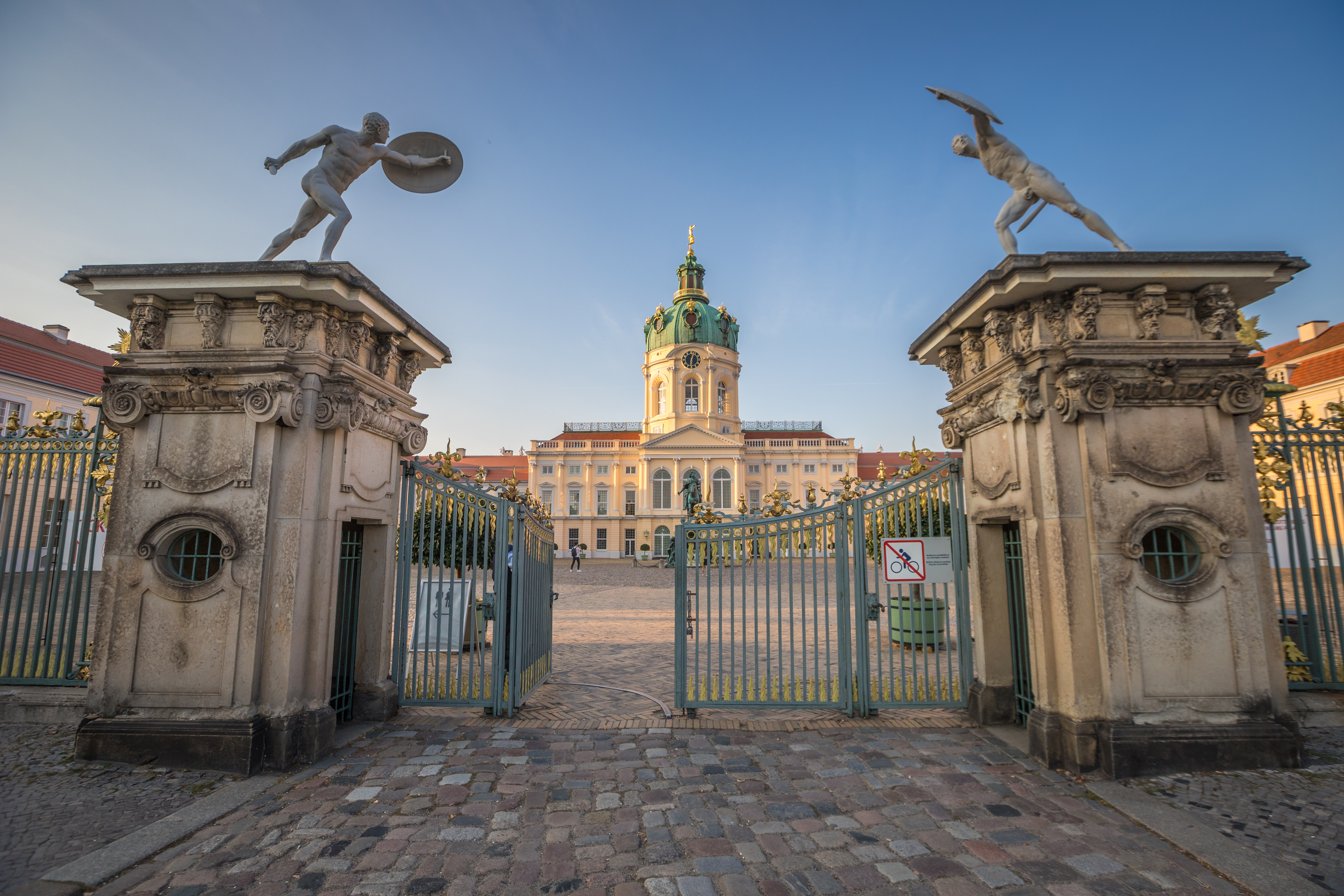 Indgangsporten til Charlottenburg Slot i Berlin med imponerende barokstatuer og slottets grønne kuppel i baggrunden