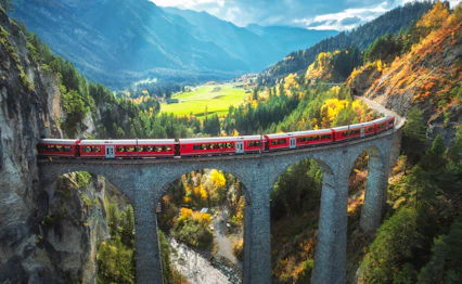 Fascinerende fugleperspektiv af det ikoniske røde Bernina Express tog på Landwasser Viadukten i Schweiz, med de majestætiske alper og flammende efterårsskove i baggrunden