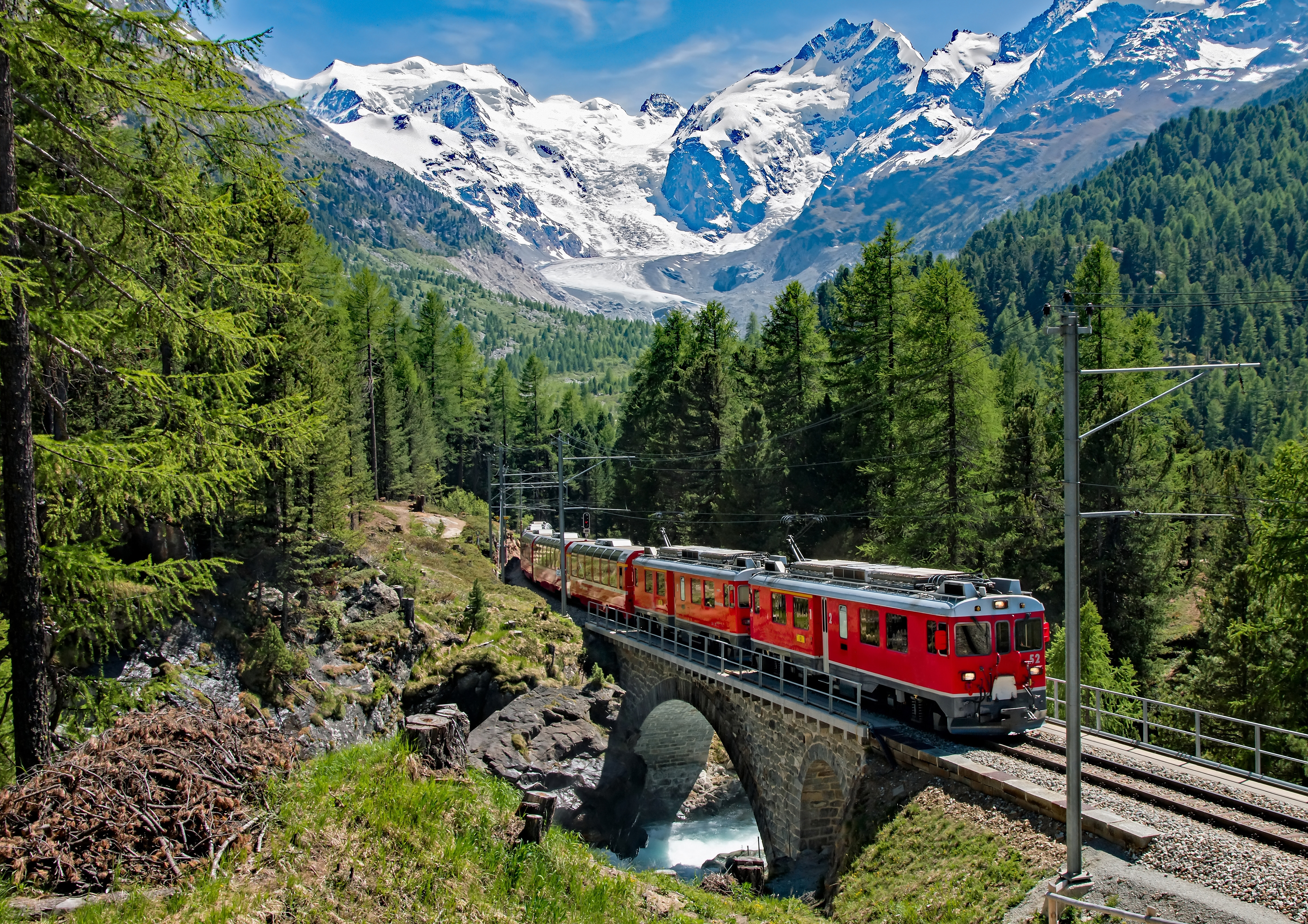 Panoramaudsigt over det røde Bernina Express tog på stenbro omgivet af grønne nåletræer og dramatiske schweiziske alper