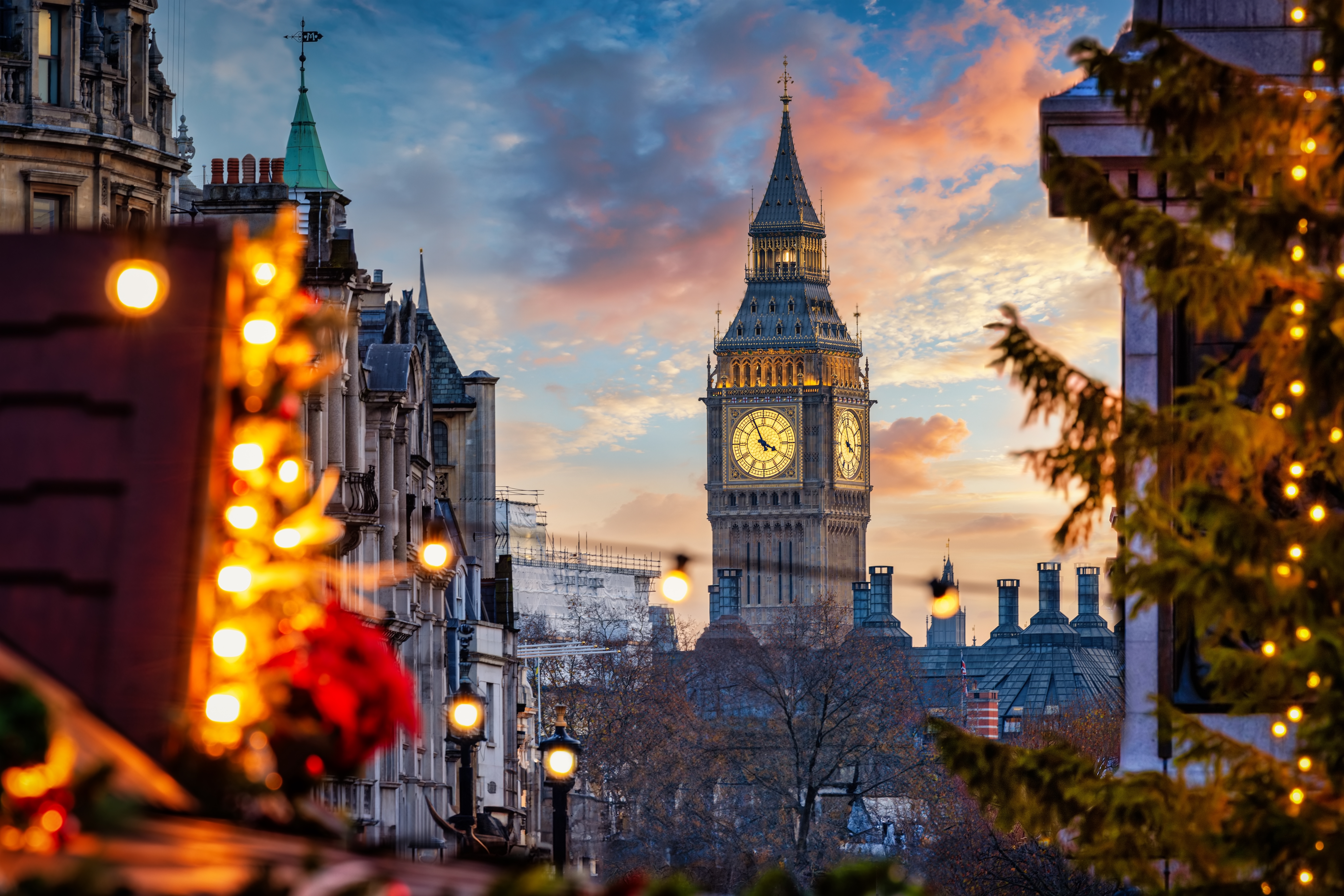 Ikonisk Big Ben klokketårn i London oplyst ved vintersonedgang med glimtende julelys fra det stemningsfulde Trafalgar Square julemarked