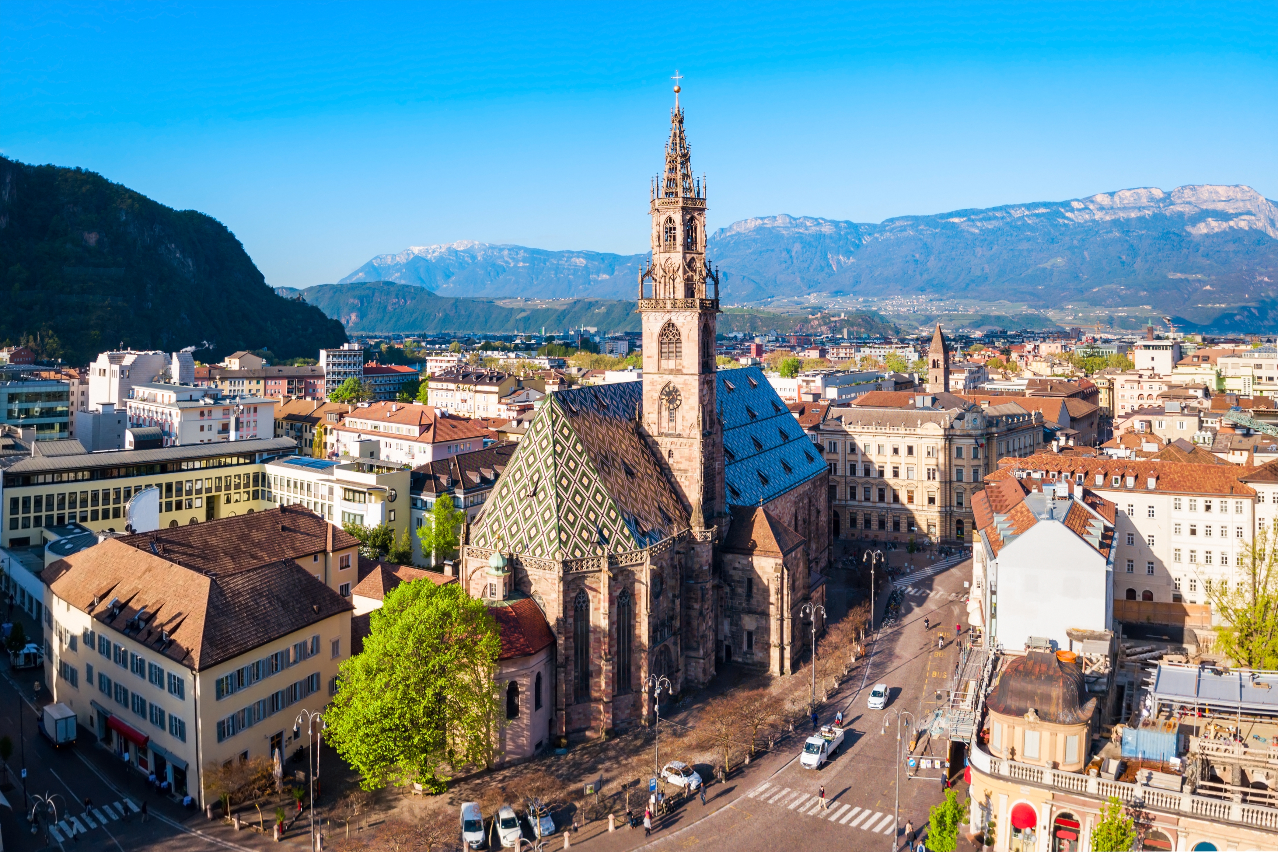 Imponerende luftfoto af den gotiske domkirke i Bolzano med det iøjnefaldende diamantmønstrede tag, omgivet af charmerende bygninger og Alperne i Sydtyrol
