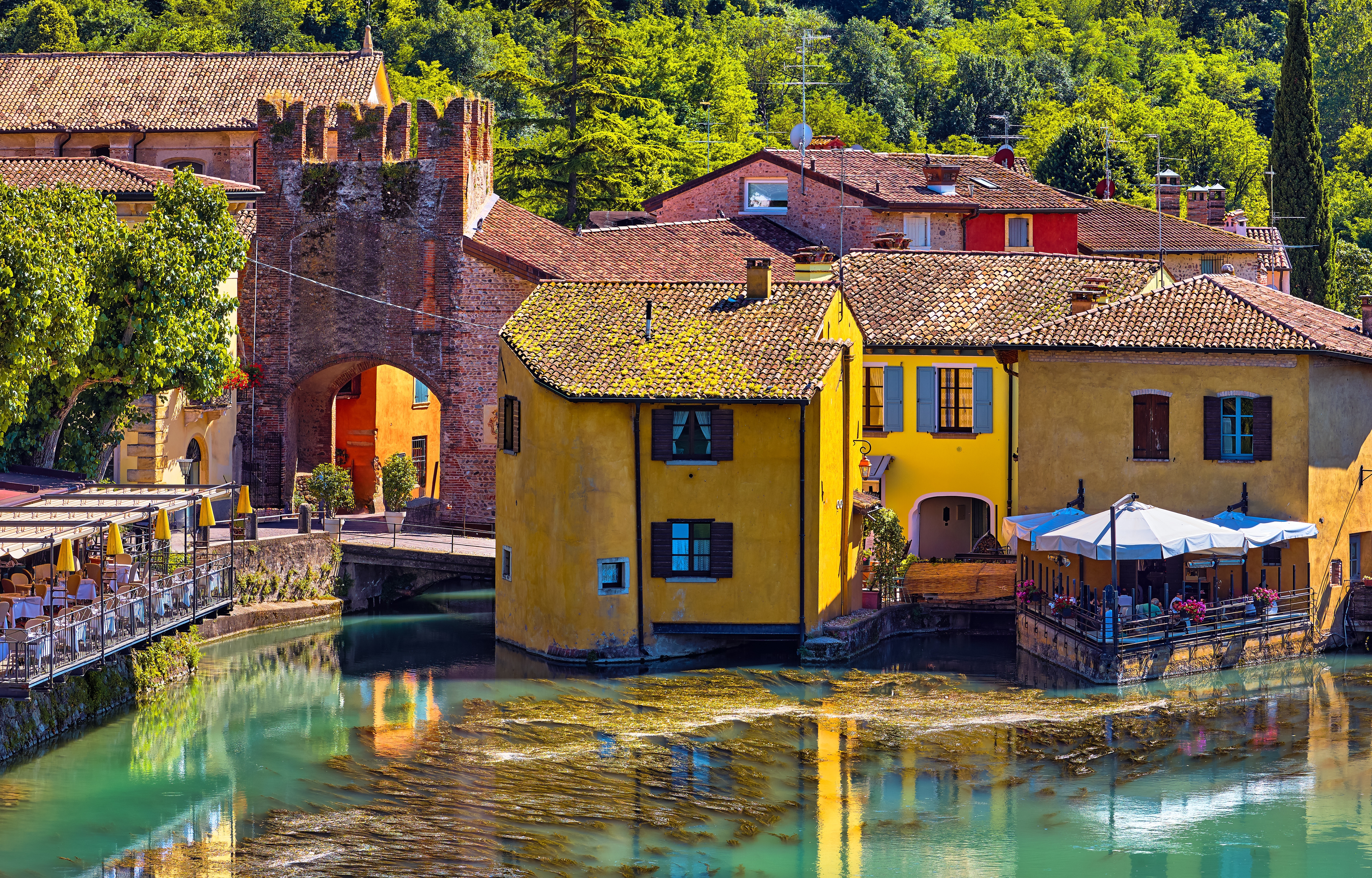 Stemningsfuld udsigt over Borghetto sul Mincio med karakteristiske gule huse ved flodbredden, hyggelige restauranter og gammel stenbue omgivet af grøn natur i Italien