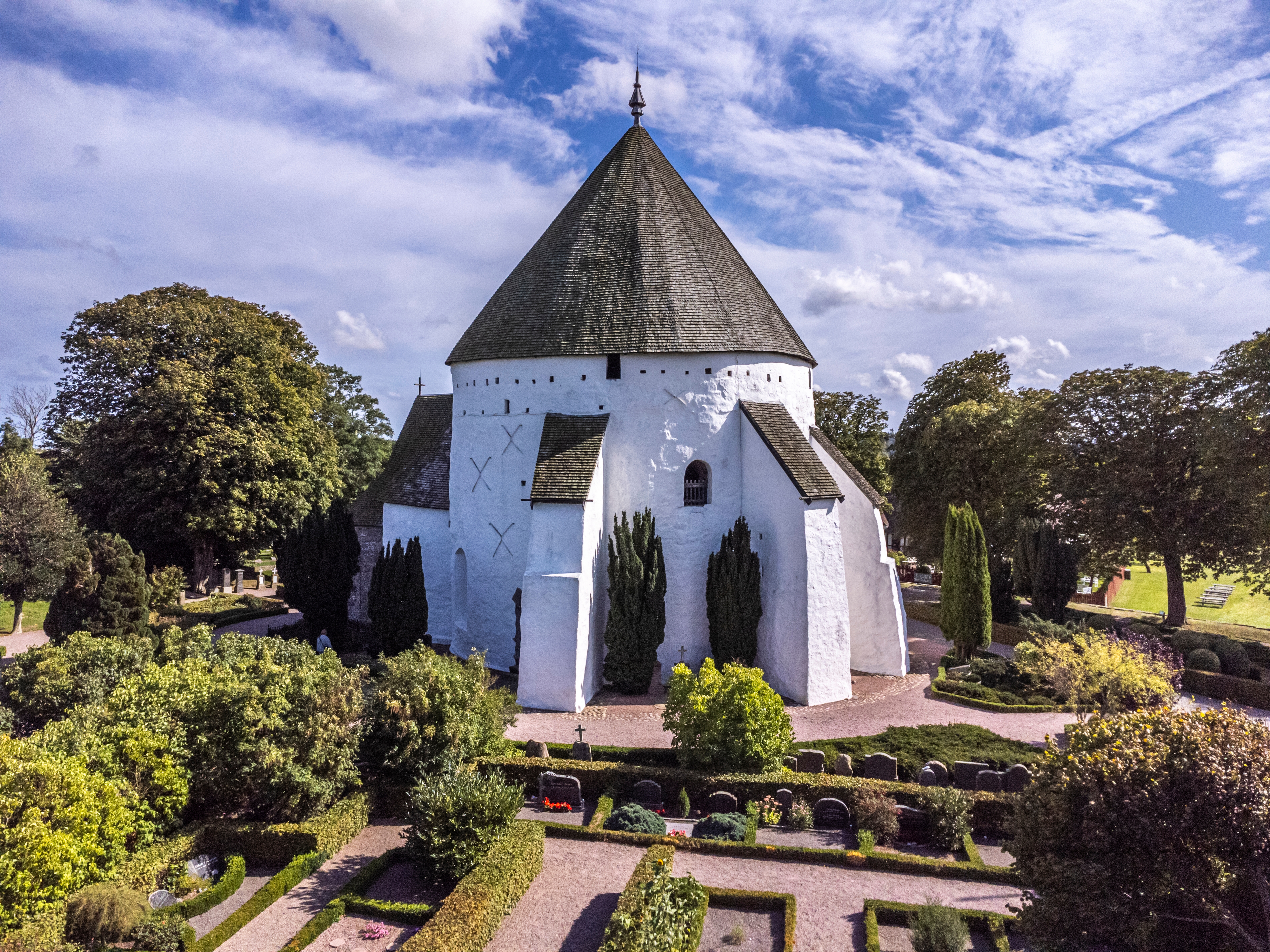 Imponerende luftfoto af den middelalderlige Østerlars Rundkirke på Bornholm med karakteristisk hvid facade og konisk tag omgivet af kirkegård og grønne træer