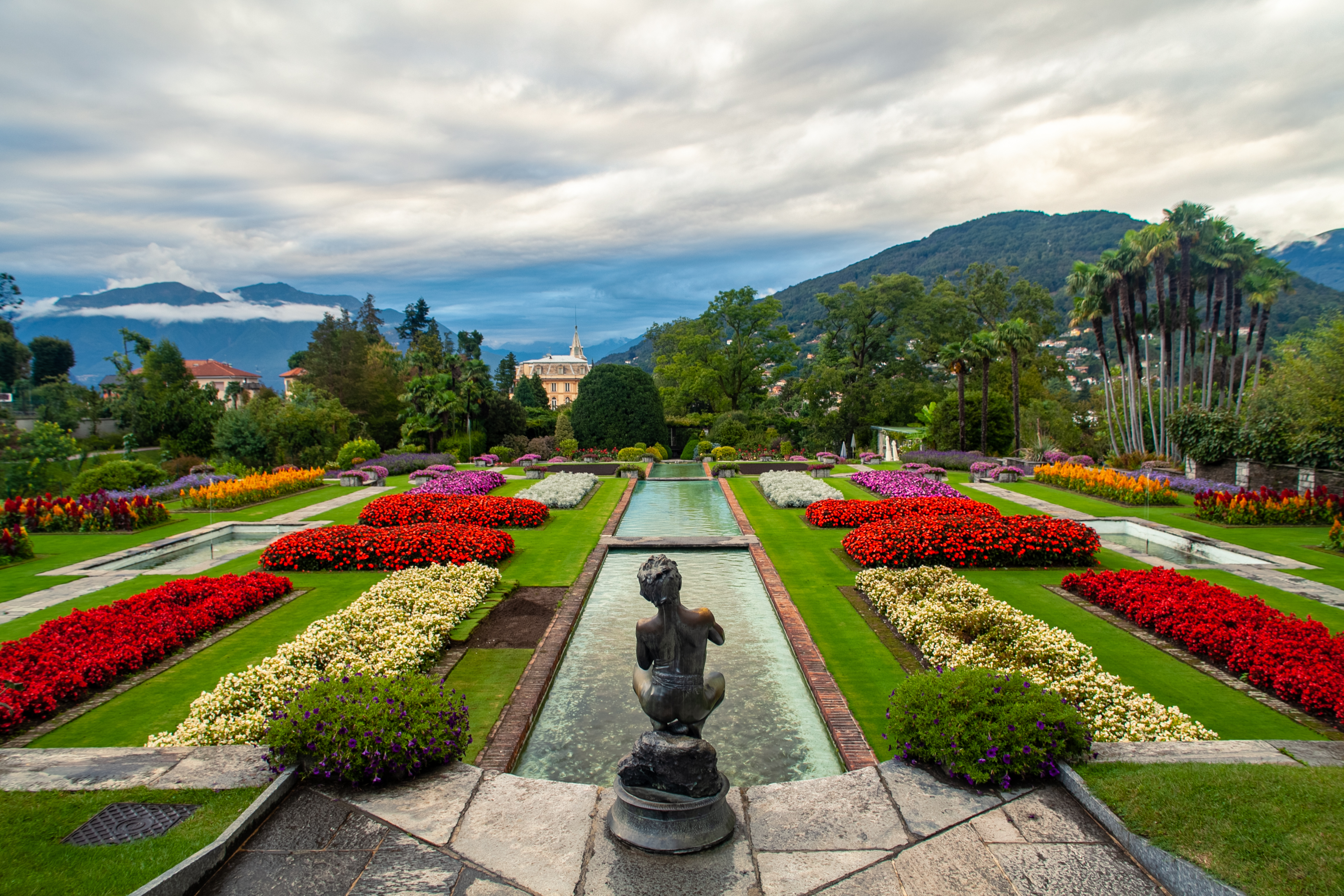 Velplejet botanisk have med røde og hvide blomster omkring vandkanal og skulptur i Villa Taranto, Piemonte med bjerglandskab i baggrunden