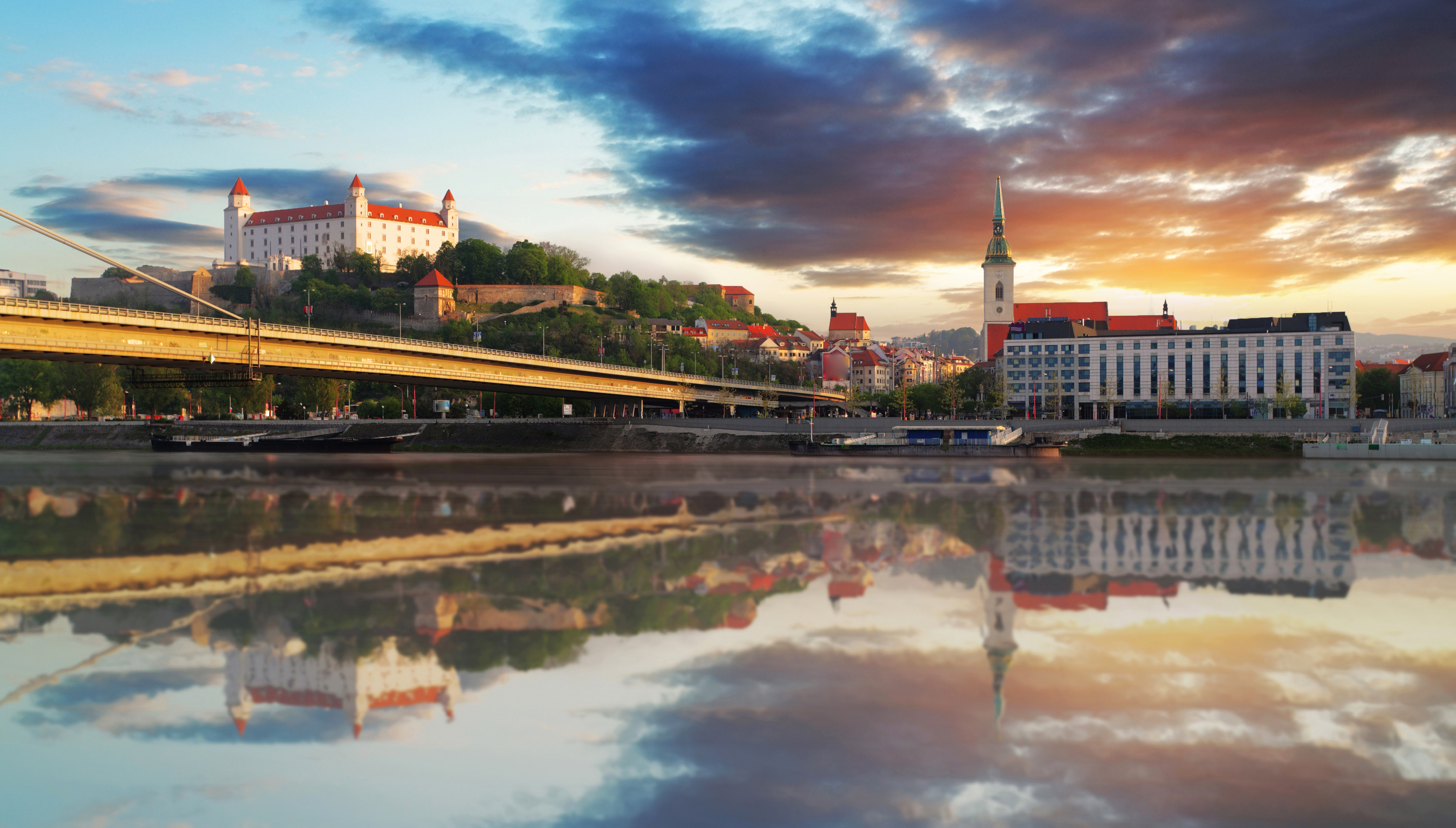 Smukt panorama over Bratislava med slottet på bakken og katedralens spir spejlet i Donau-floden