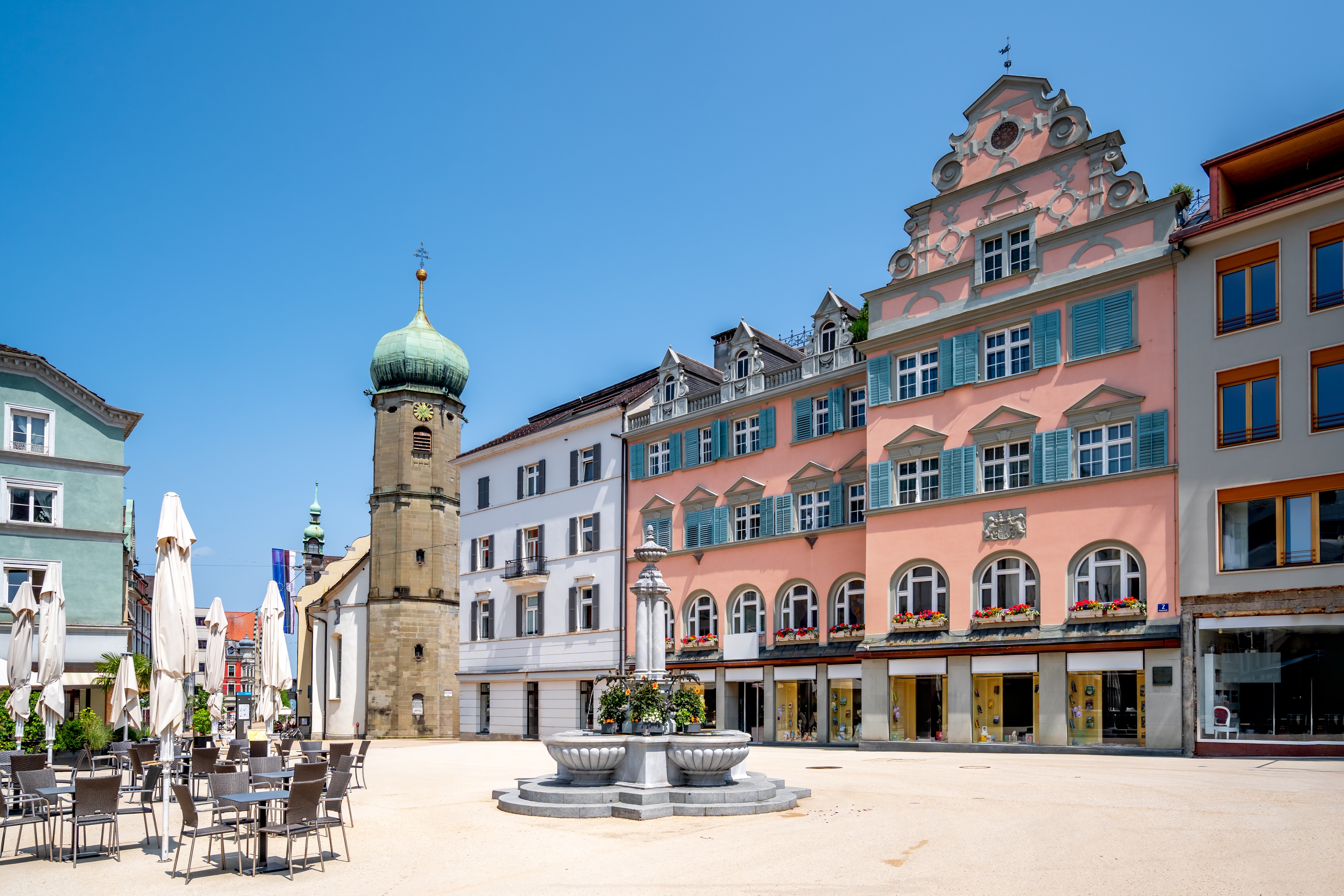 Idyllisk torv i Bregenz med farverige historiske bygninger, dekorativt springvand og café-miljø under blå himmel i Østrig