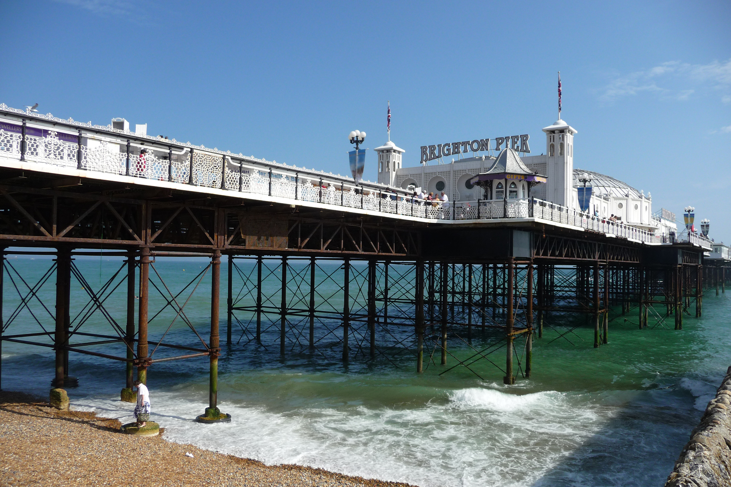 Brighton Pier med sin karakteristiske arkitektur og bølger der slår mod søjlerne på en solrig sommerdag ved den engelske kyst