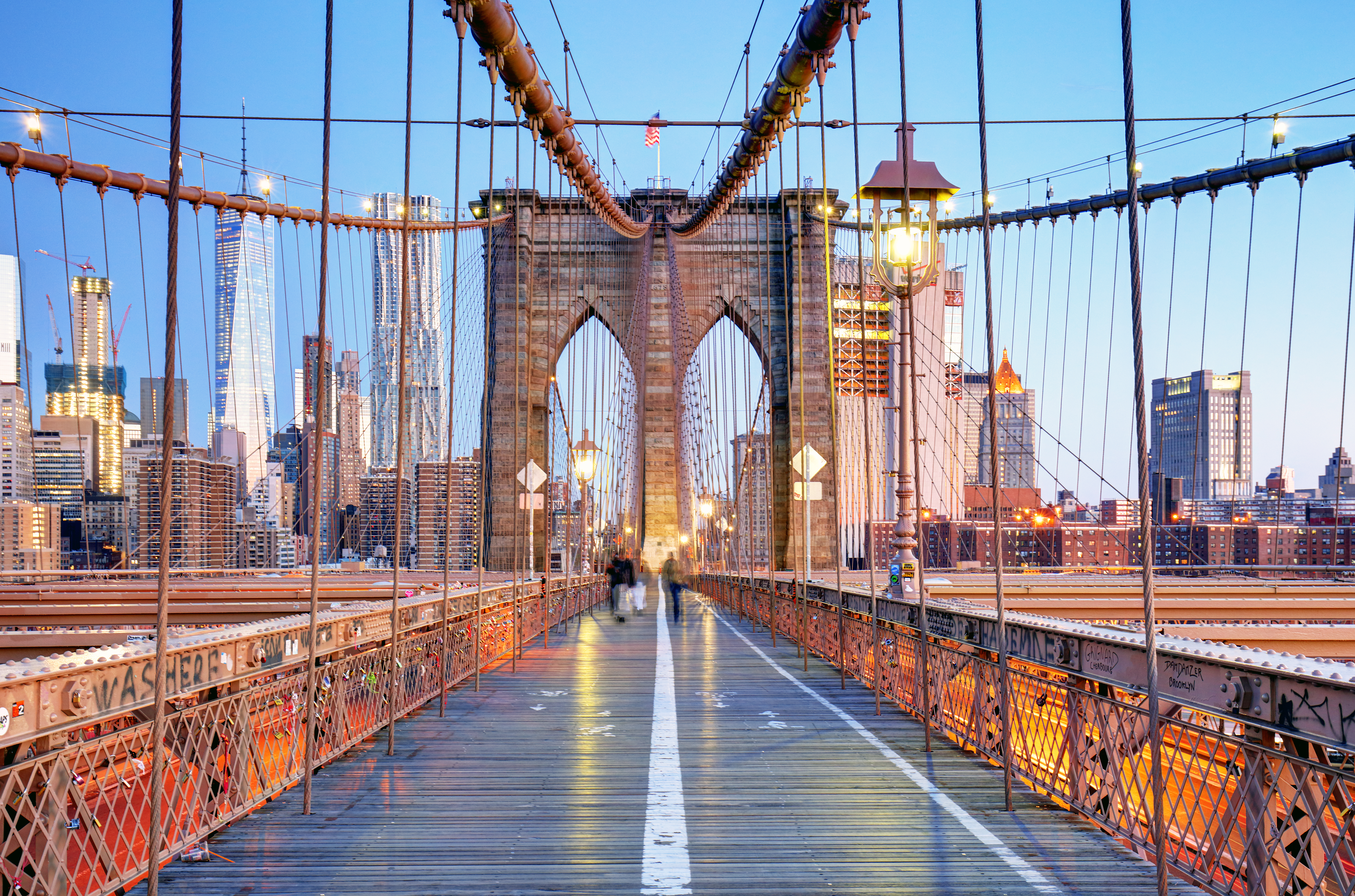 Skumringsvy langs Brooklyn Bridge med majestætiske buer og kabler, der strækker sig mod Manhattans lysende skyskrabere. East River glimter nedenfor i dette ikoniske New York-panorama.