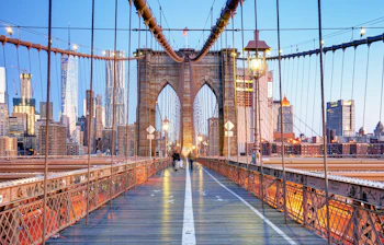 Skumringsvy langs Brooklyn Bridge med majestætiske buer og kabler, der strækker sig mod Manhattans lysende skyskrabere. East River glimter nedenfor i dette ikoniske New York-panorama.
