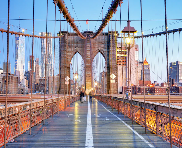Skumringsvy langs Brooklyn Bridge med majestætiske buer og kabler, der strækker sig mod Manhattans lysende skyskrabere. East River glimter nedenfor i dette ikoniske New York-panorama.