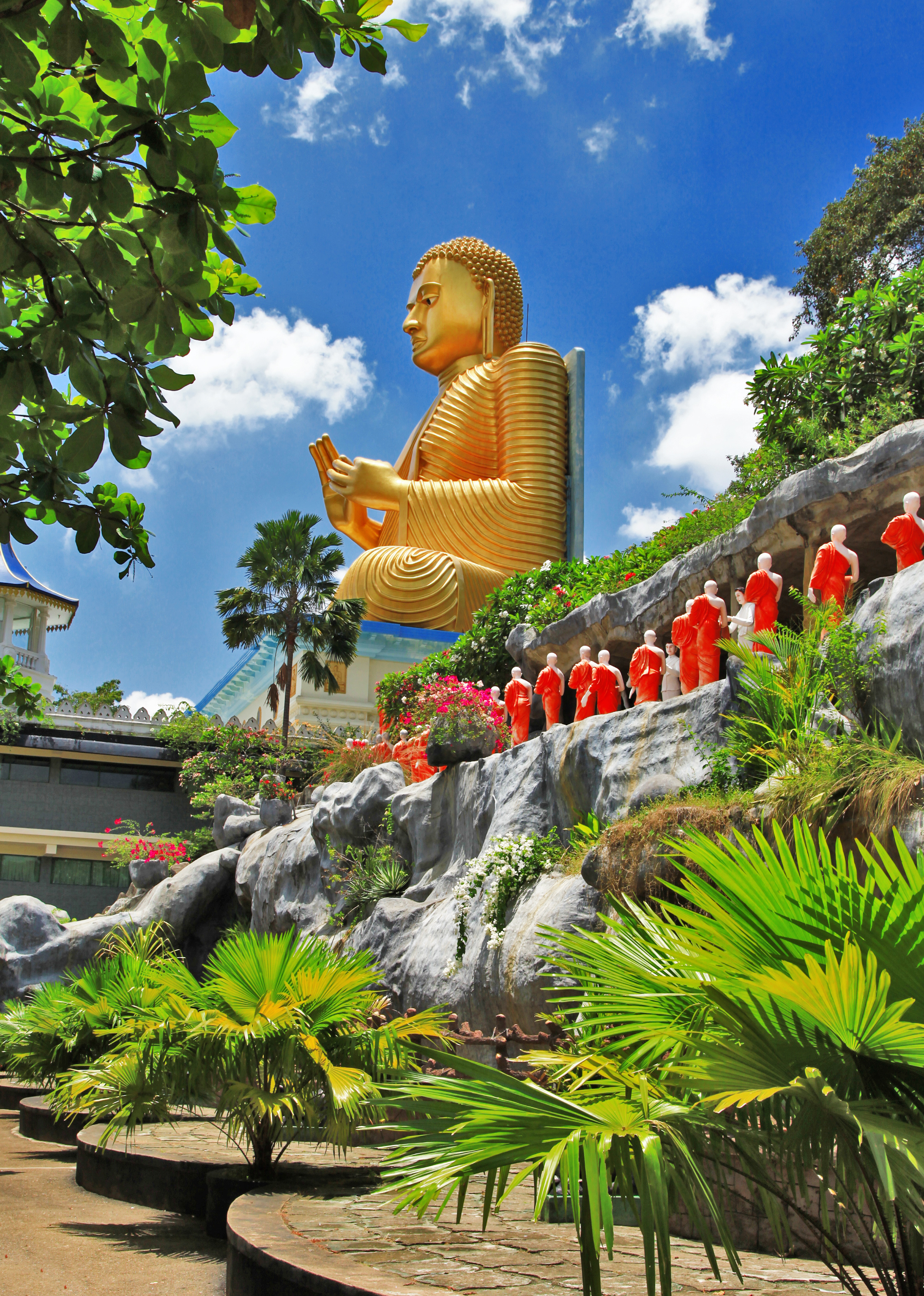 Smuk gyldne Buddha-statue ved det UNESCO-listede Dambulla-tempel i Sri Lanka omgivet af tropisk natur og oransjerøde munkefigurer