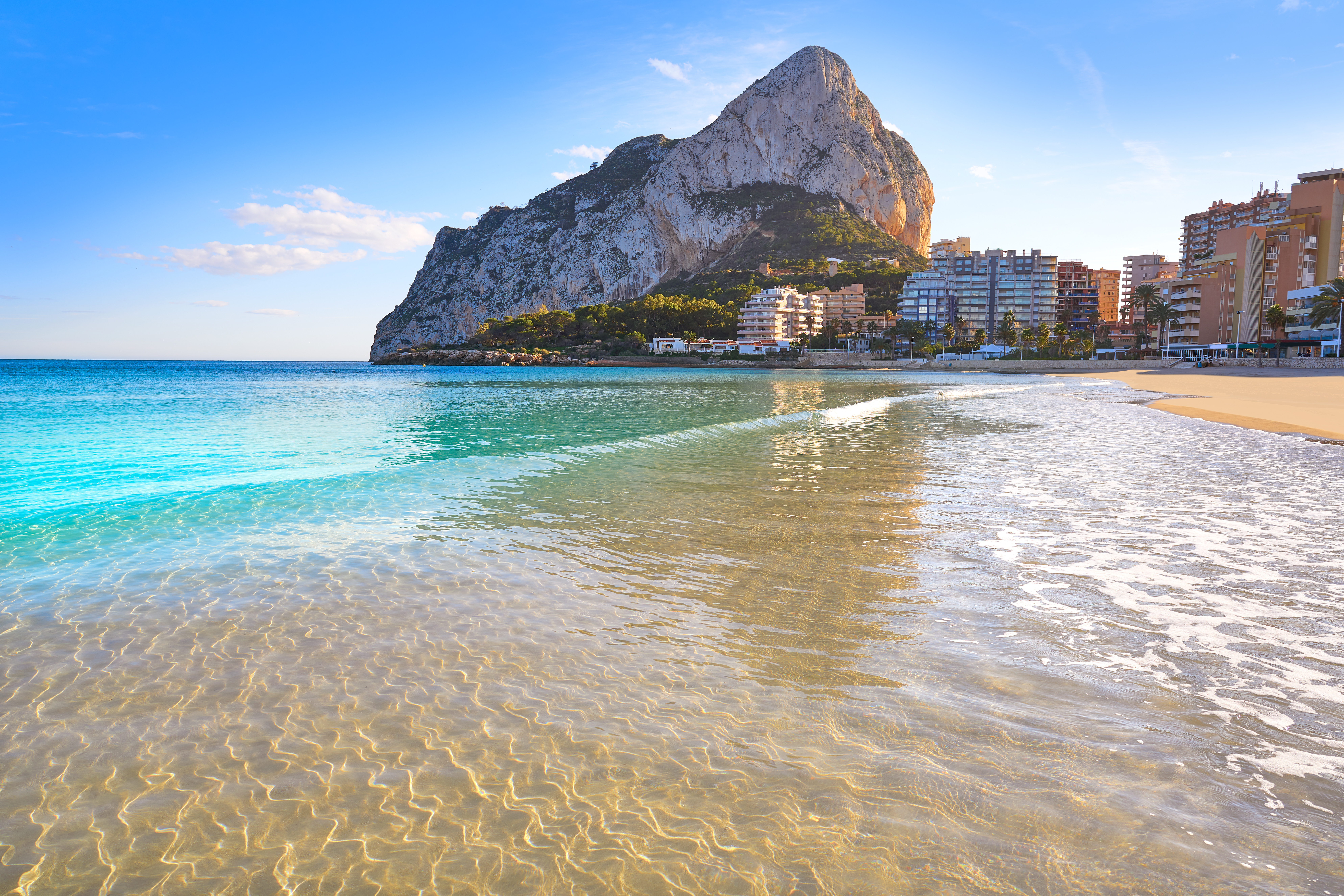 Idyllisk sandstrand i Calpe ved Alicante med det kendte Penon de Ifach klippemassiv og klart turkisblåt Middelhavsvand under blå himmel