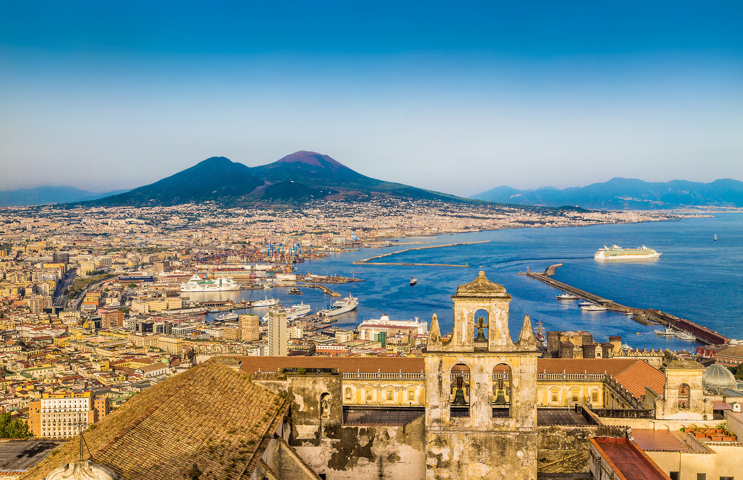 Storslået panorama over Napoli i aftenlys med den karakteristiske Vesuv-vulkan, der rejser sig bag byens udstrakte bebyggelse langs den smukke Middelhavskyst i Campania-regionen