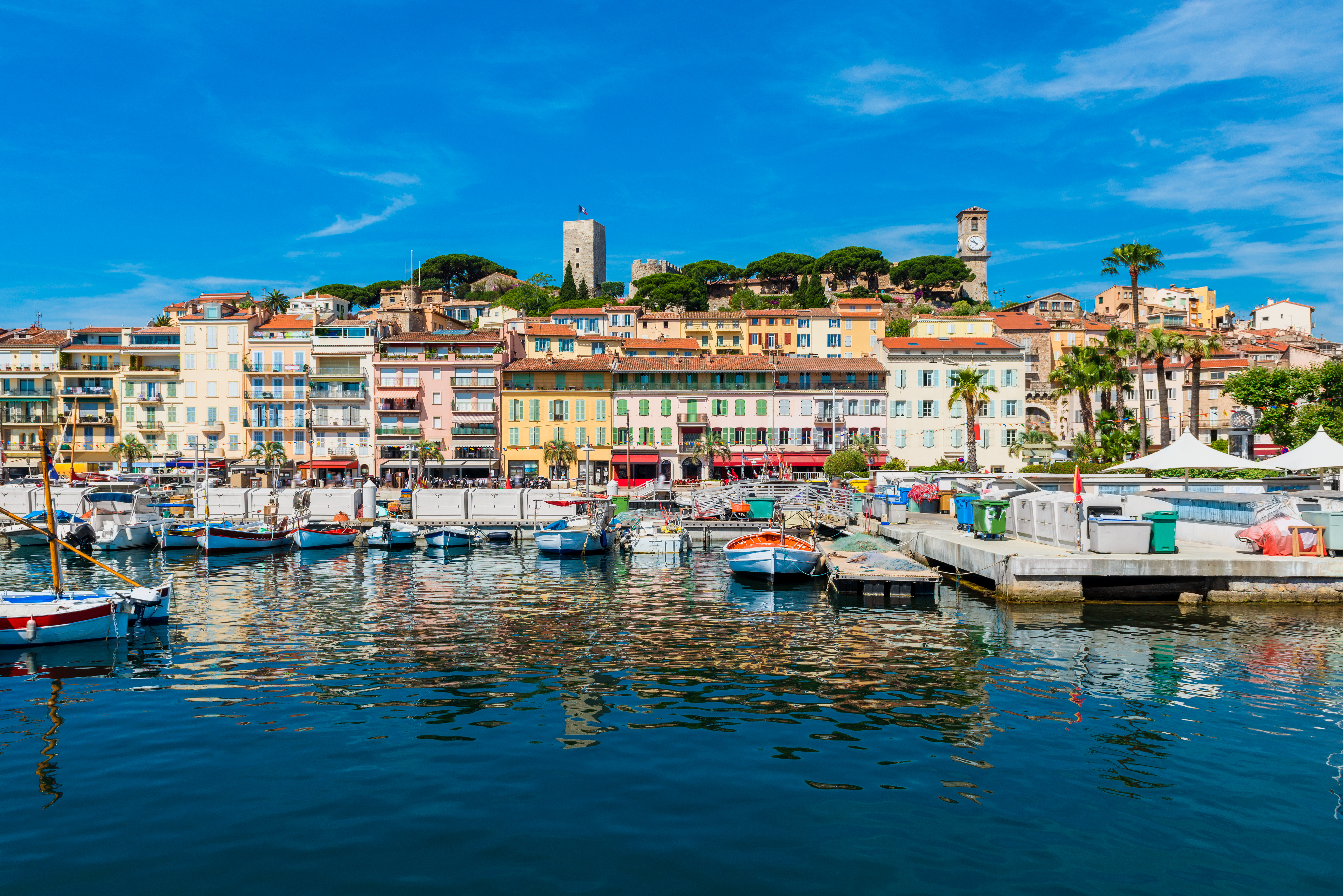 Idyllisk udsigt over Cannes havn med det historiske Le Suquet-kvarter, farverige bygninger langs kajkanten og små både der ligger fortøjet på en solrig sommerdag
