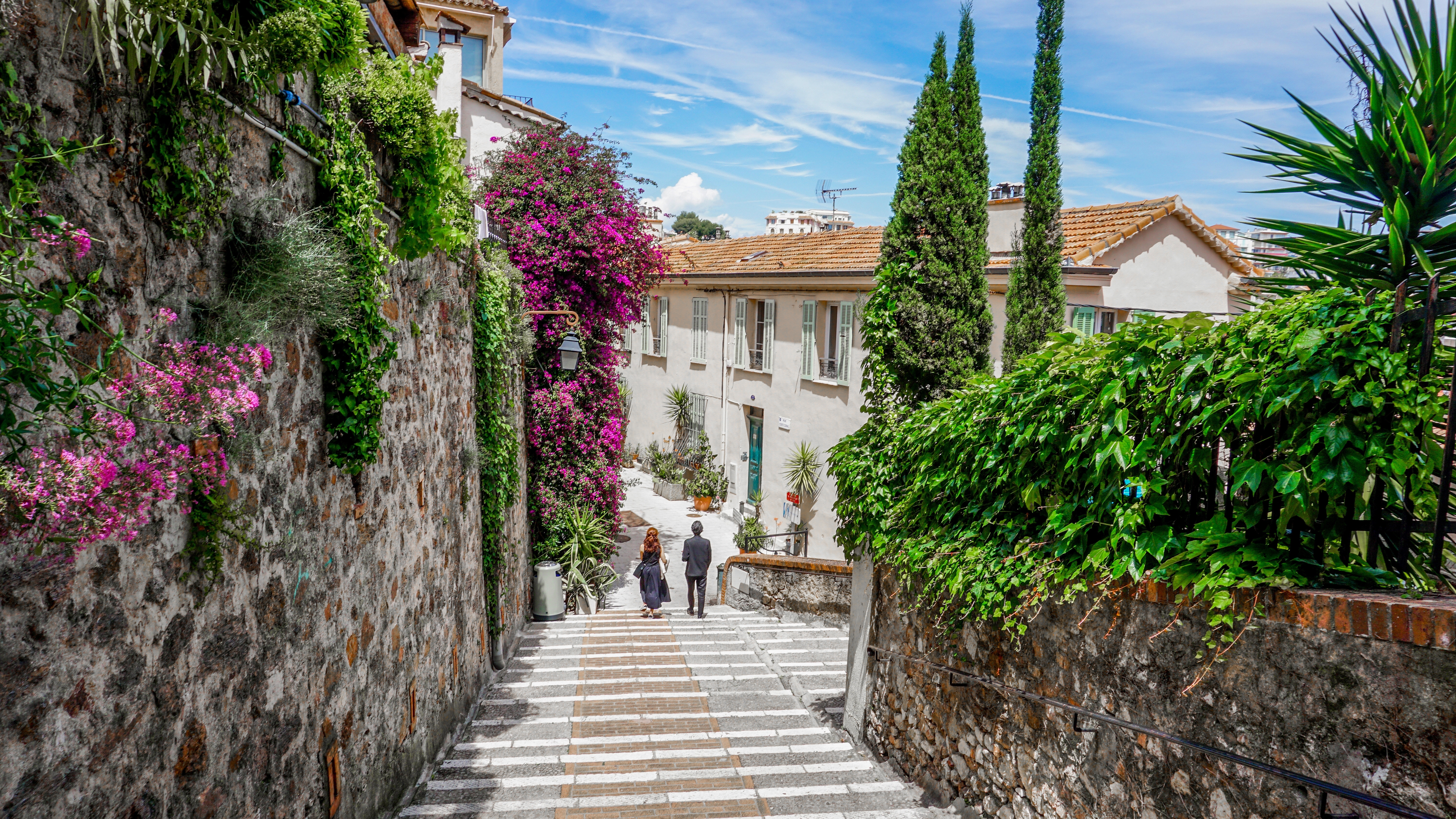 Par går ned ad en charmerende stentrappe i Cannes gamle bydel med smukke lyserøde blomster og frodig grøn vegetation