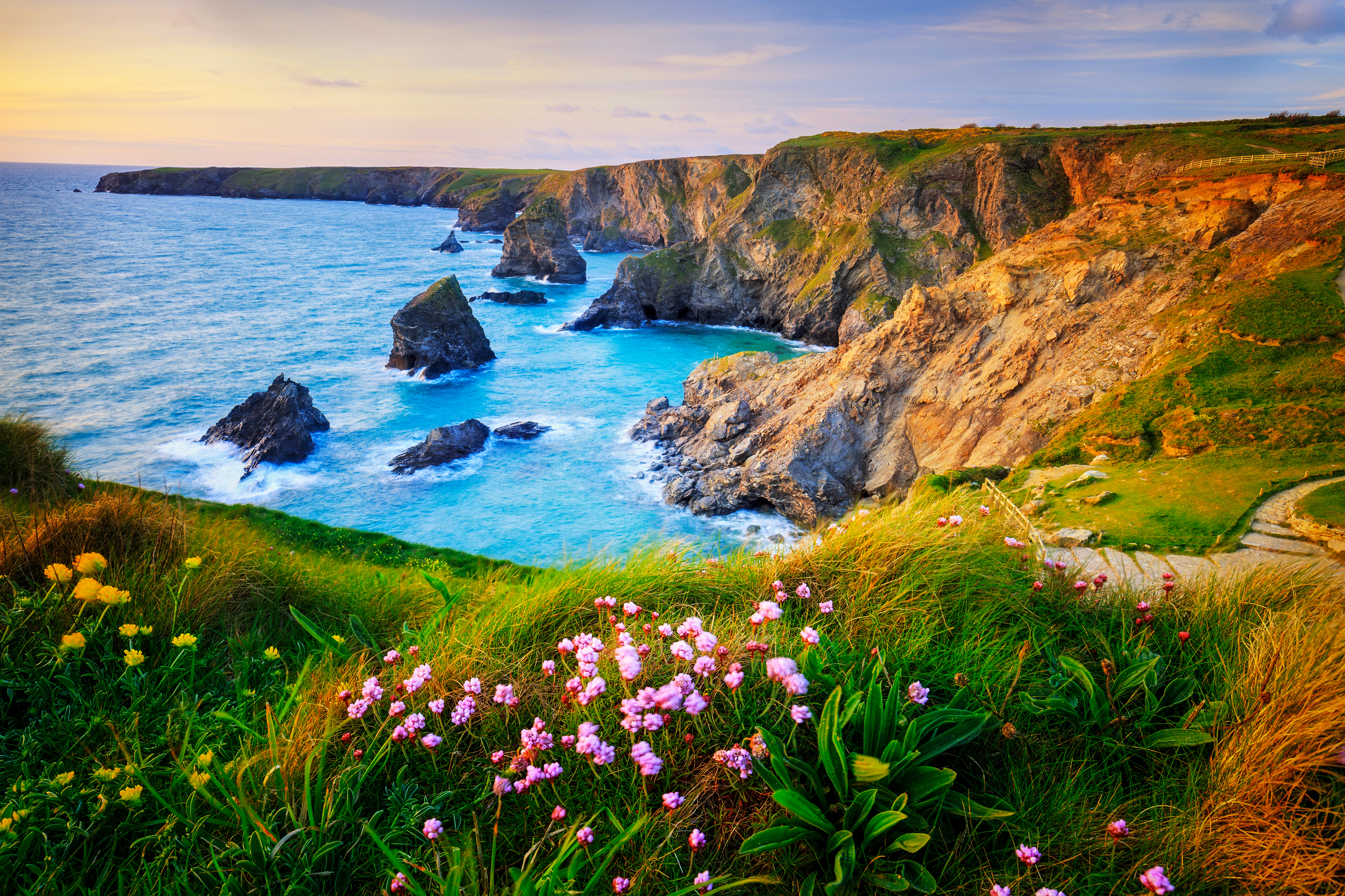 Naturskønt kystparti ved Bedruthan Steps i Cornwall, England, hvor solnedgangens gyldne lys fremhæver de dramatiske klipper, azurblåt hav og blomstrende vegetation