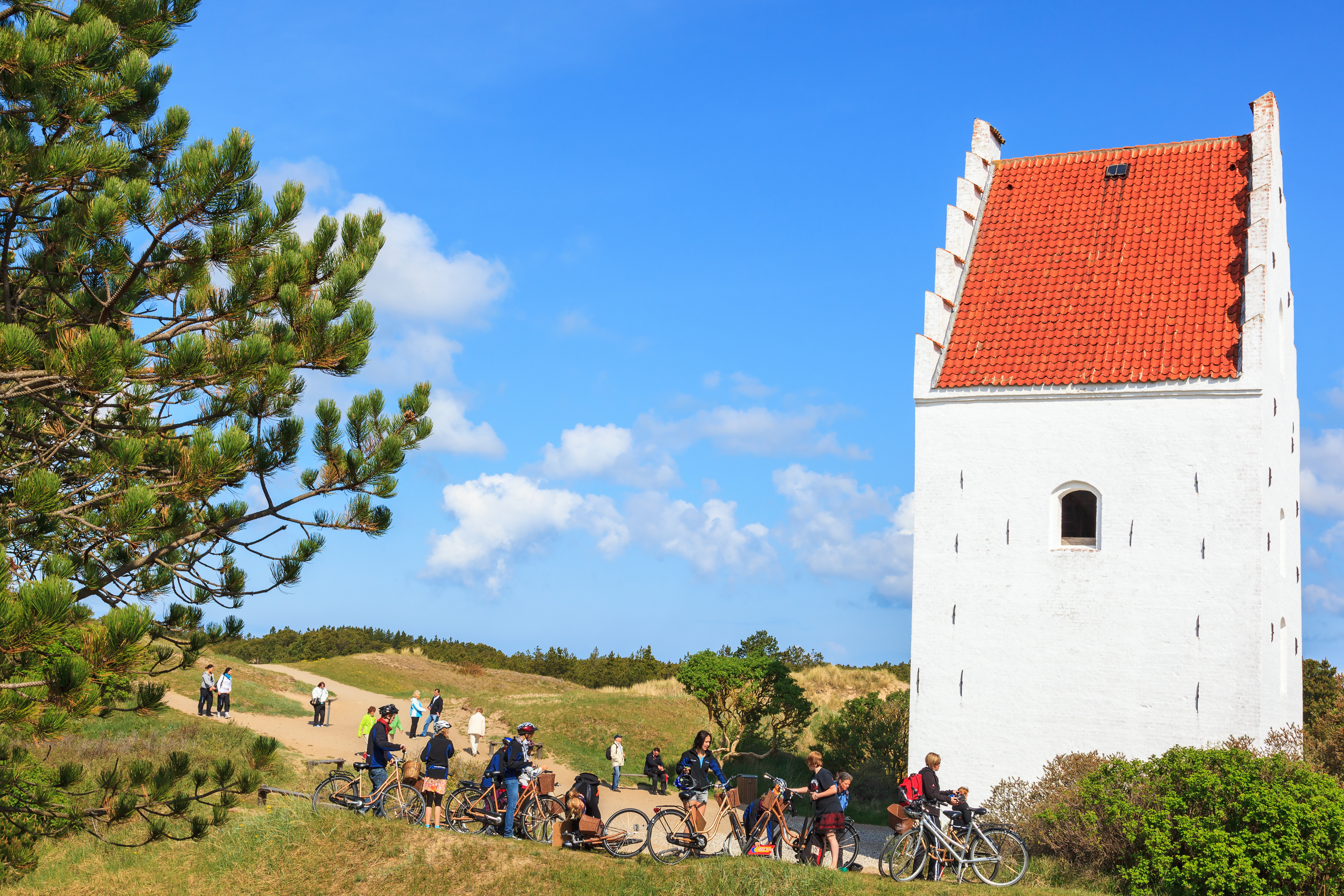 Cyklister på tur ved den berømte Tilsandede Kirke i Skagen med det hvide kirketårn og røde tag mod blå himmel