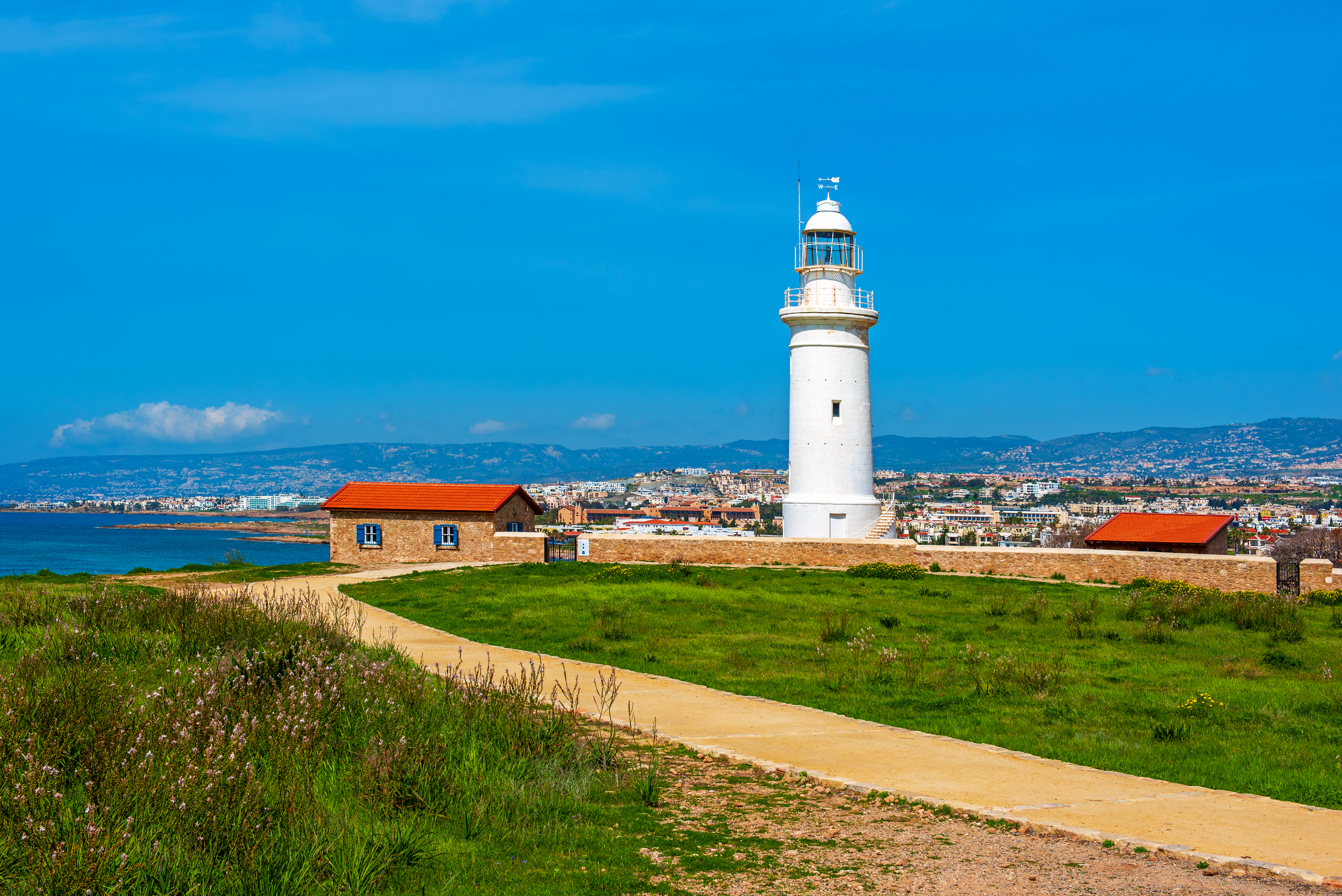Fyrtårnet ved Paphos arkæologiske park på Cypern med grøn sti og blåt hav under en klar sommerblå himmel