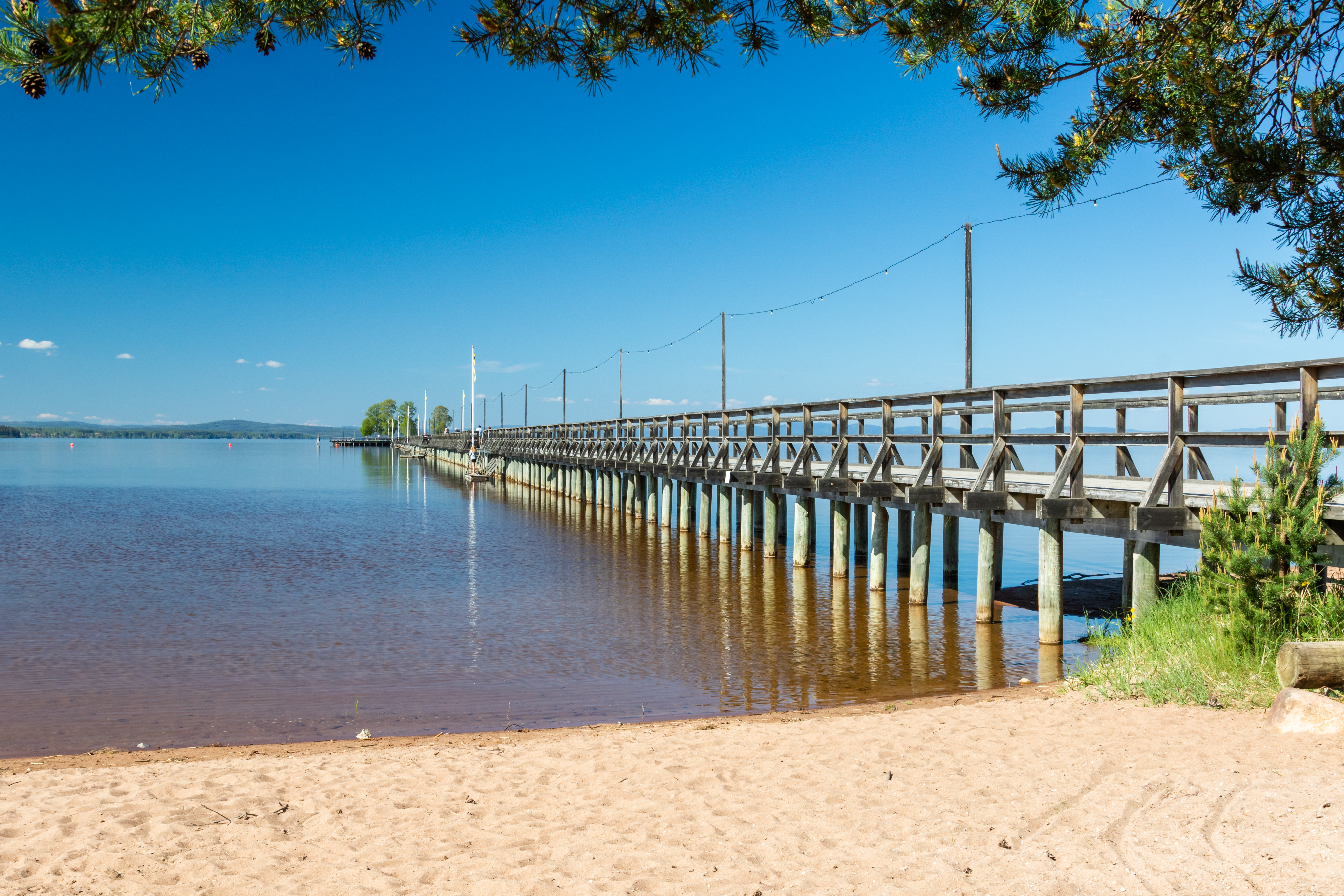 Naturskøn træbro over den stille sø i Rättvik, Dalarna, Sverige, omgivet af sandstrand og grønne træer på en solrig dag