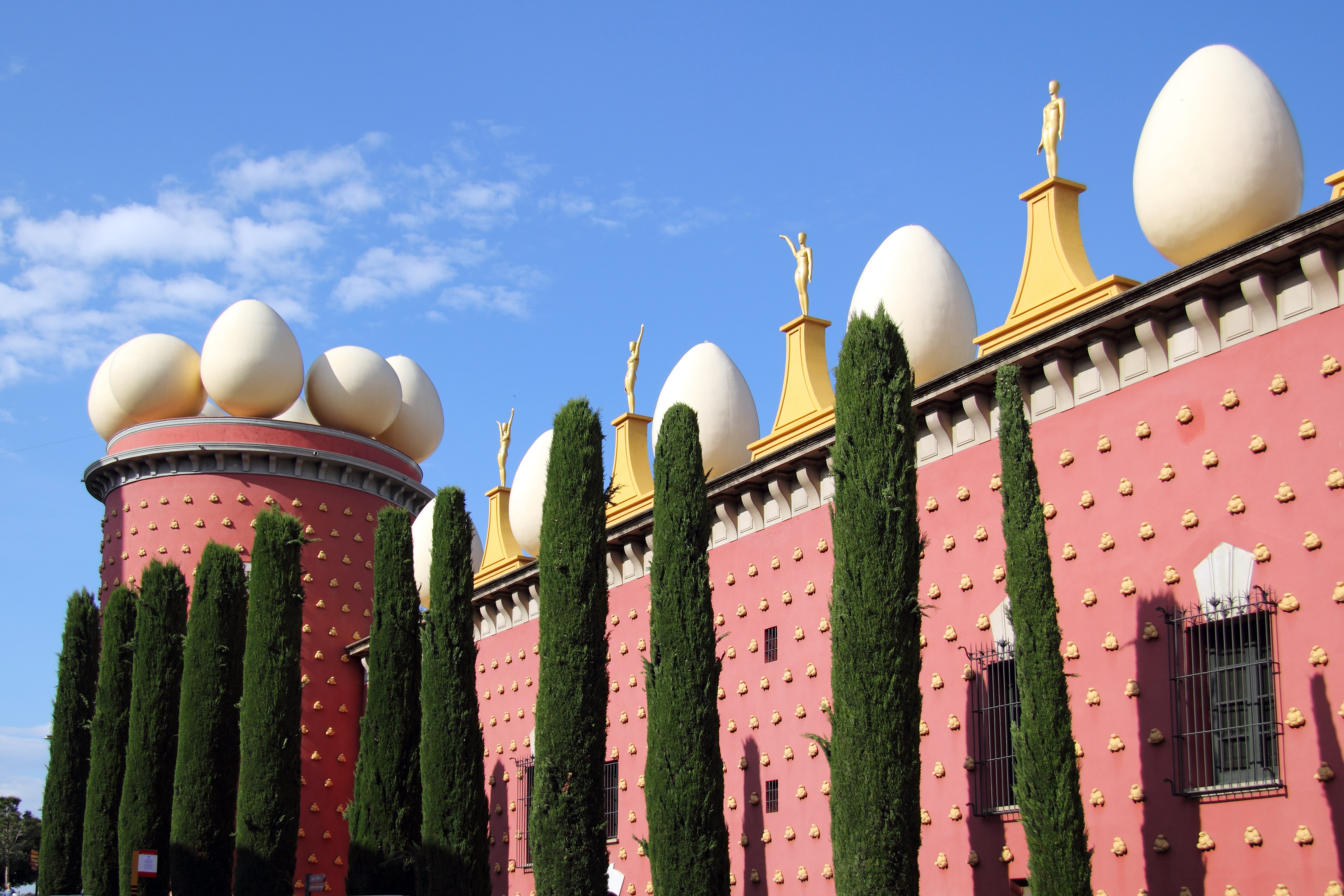 Salvador Dalí Museets unikke røde facade med surrealistiske hvide æg på taget og cyprestræer i forgrunden, beliggende i Figueres