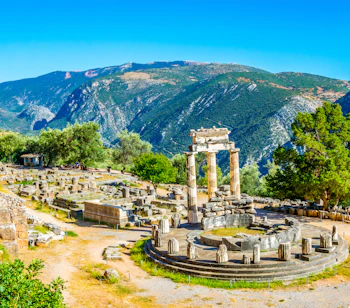Smukke antikke ruiner ved Athena Pronaia-helligdommen i Delphi, Grækenland, med tre stående søjler og UNESCO-beskyttet arkæologisk site omgivet af grønne bjerge
