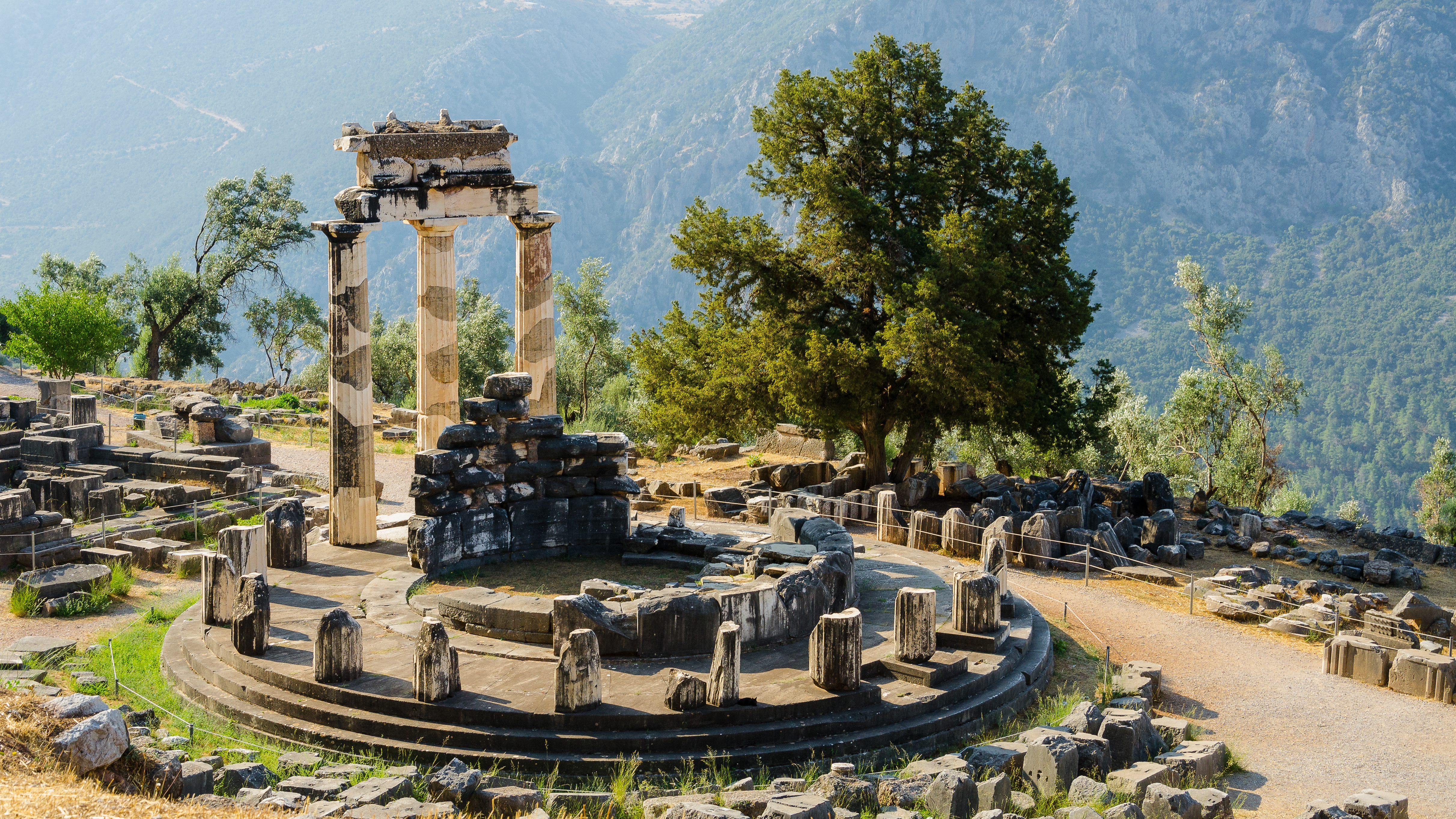Det runde Tholos-tempel i Delphi med tre bevarede søjler og spredte stenruiner foran et skovklædt bjerglandskab i Grækenland