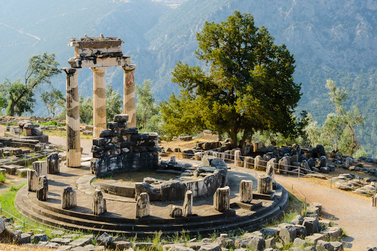 Det runde Tholos-tempel i Delphi med tre bevarede søjler og spredte stenruiner foran et skovklædt bjerglandskab i Grækenland