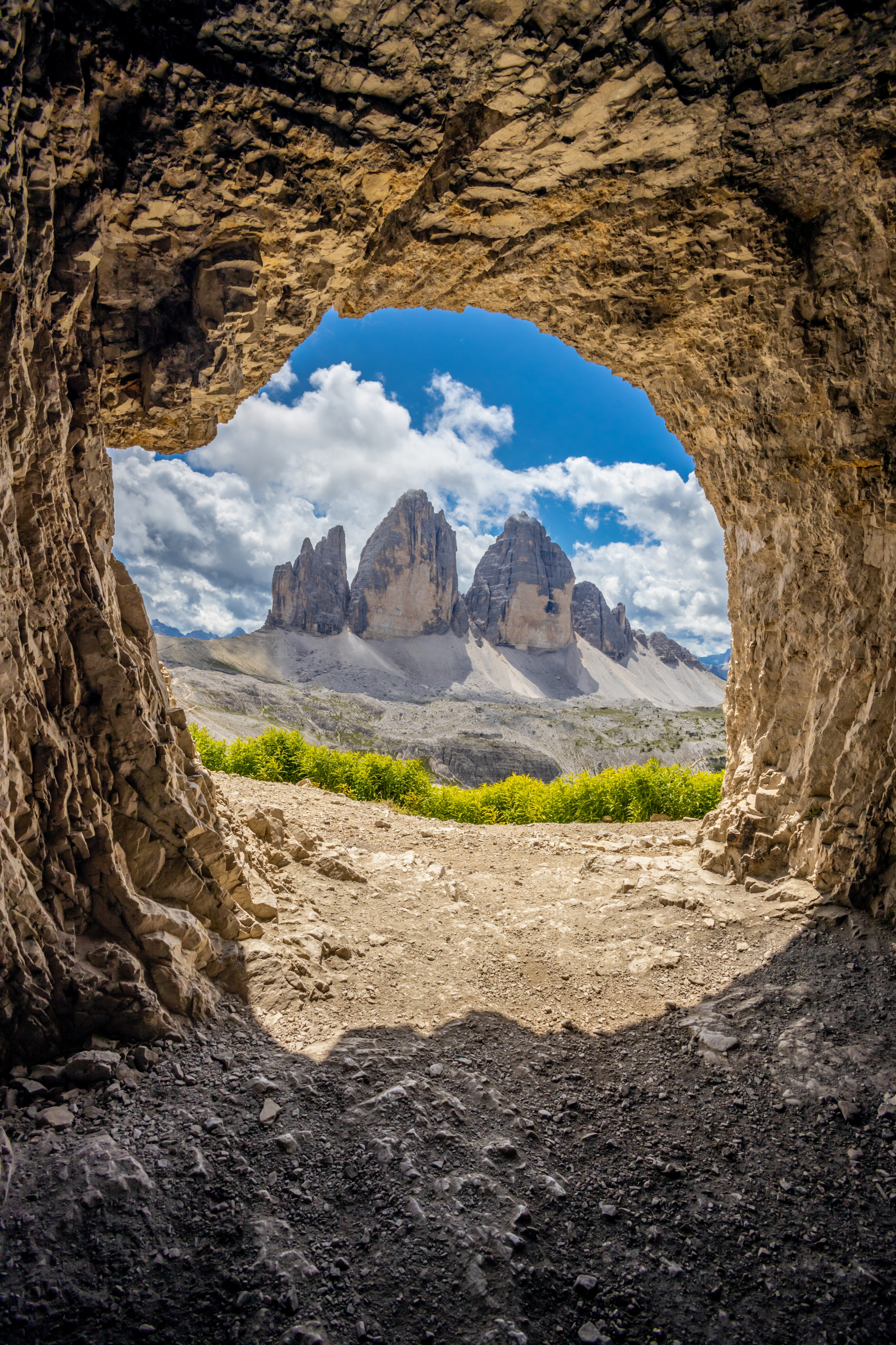Tre Cime di Lavaredo i Dolomitterne set gennem en naturlig klippehule med gule alpeblomster i forgrunden og blå sommerhimmel