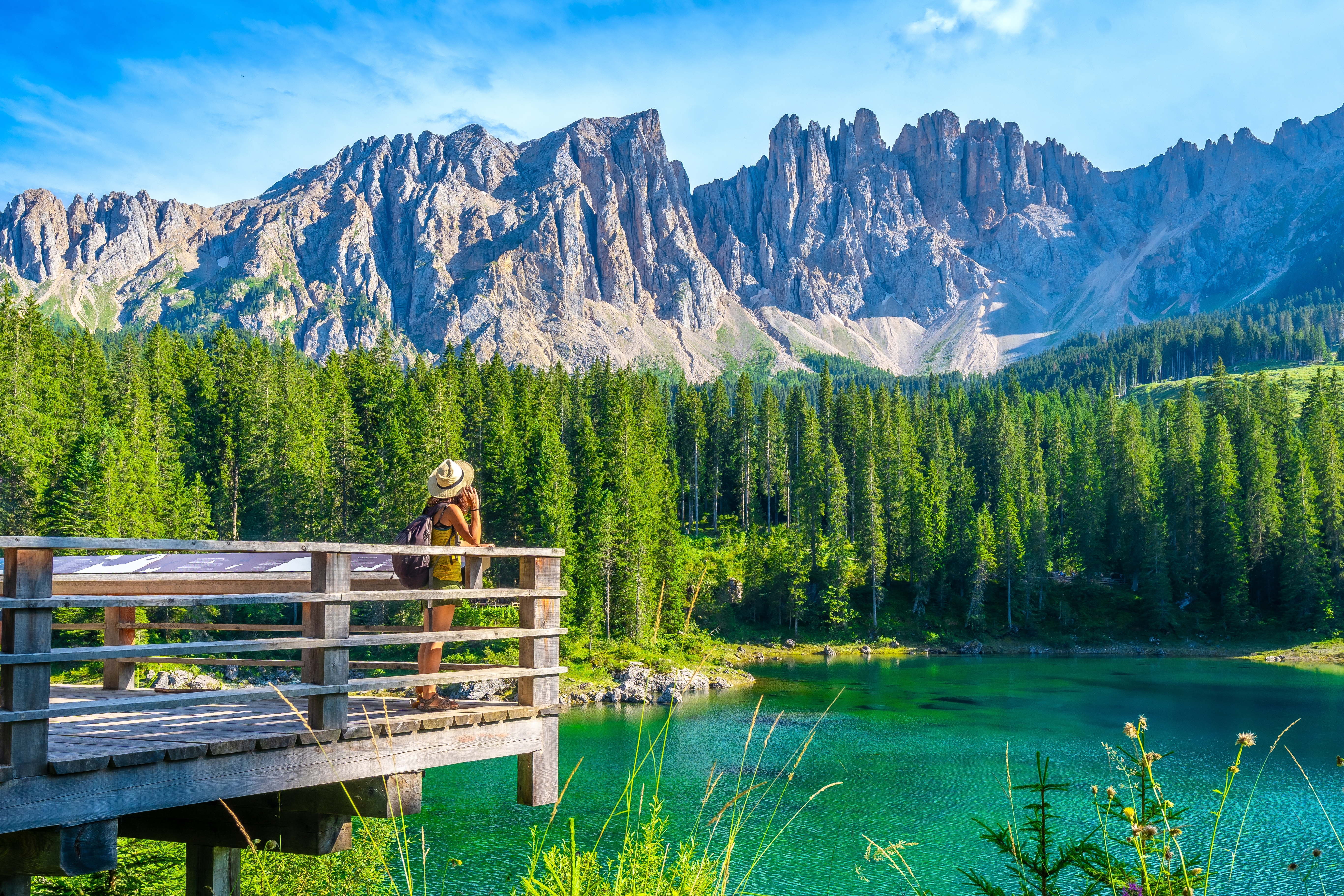 Turkisblå alpesø Lake Carezza i de italienske Dolomitter med dramatiske Latemar-bjerge og tæt grøn fyrreskov – kvindelig rejsende nyder udsigten fra træplatform om sommeren
