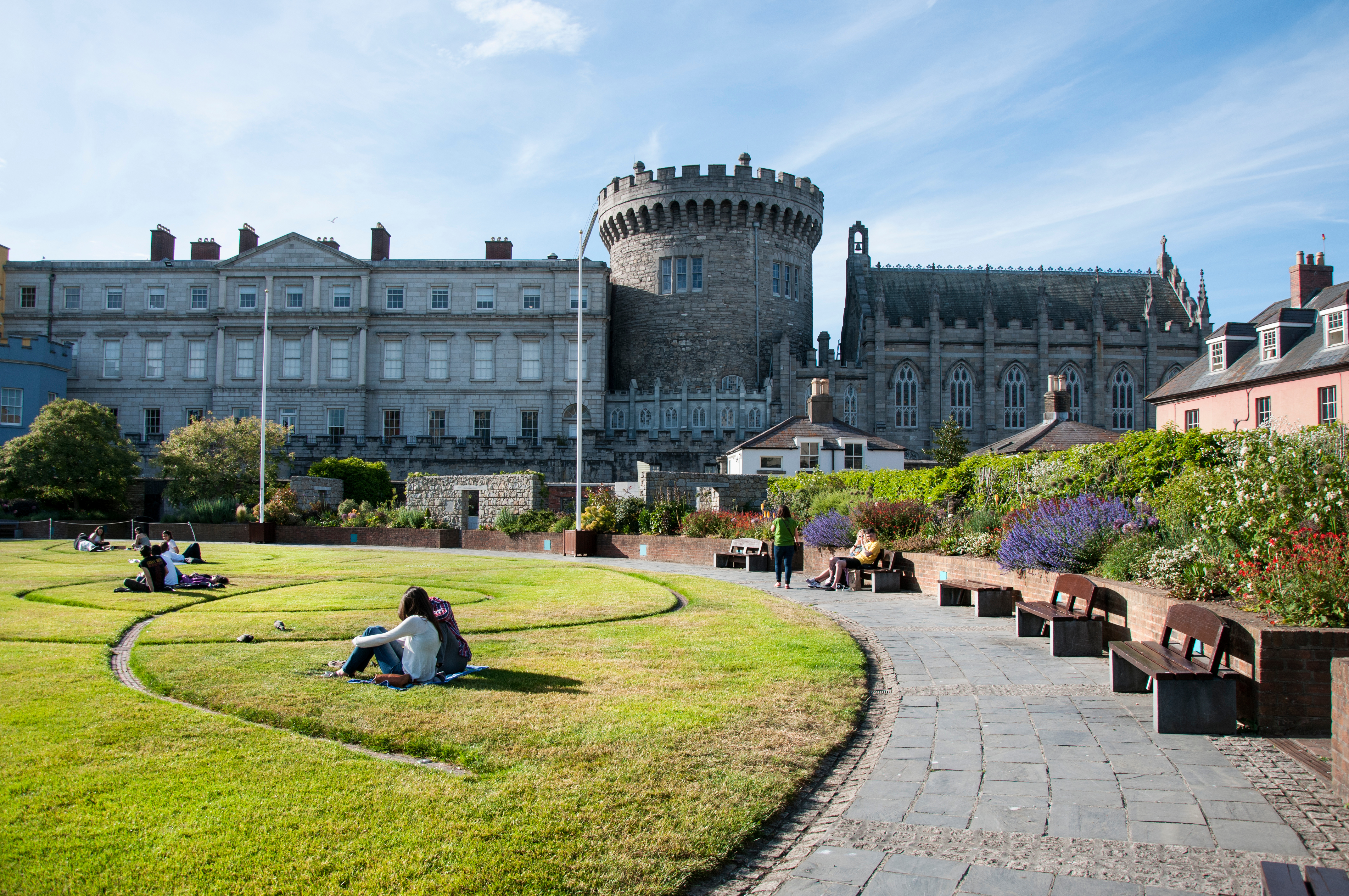 Turister nyder en solrig dag i de grønne haver ved Dublin Castle med det historiske Record Tower og slottets bygninger som dramatisk baggrund