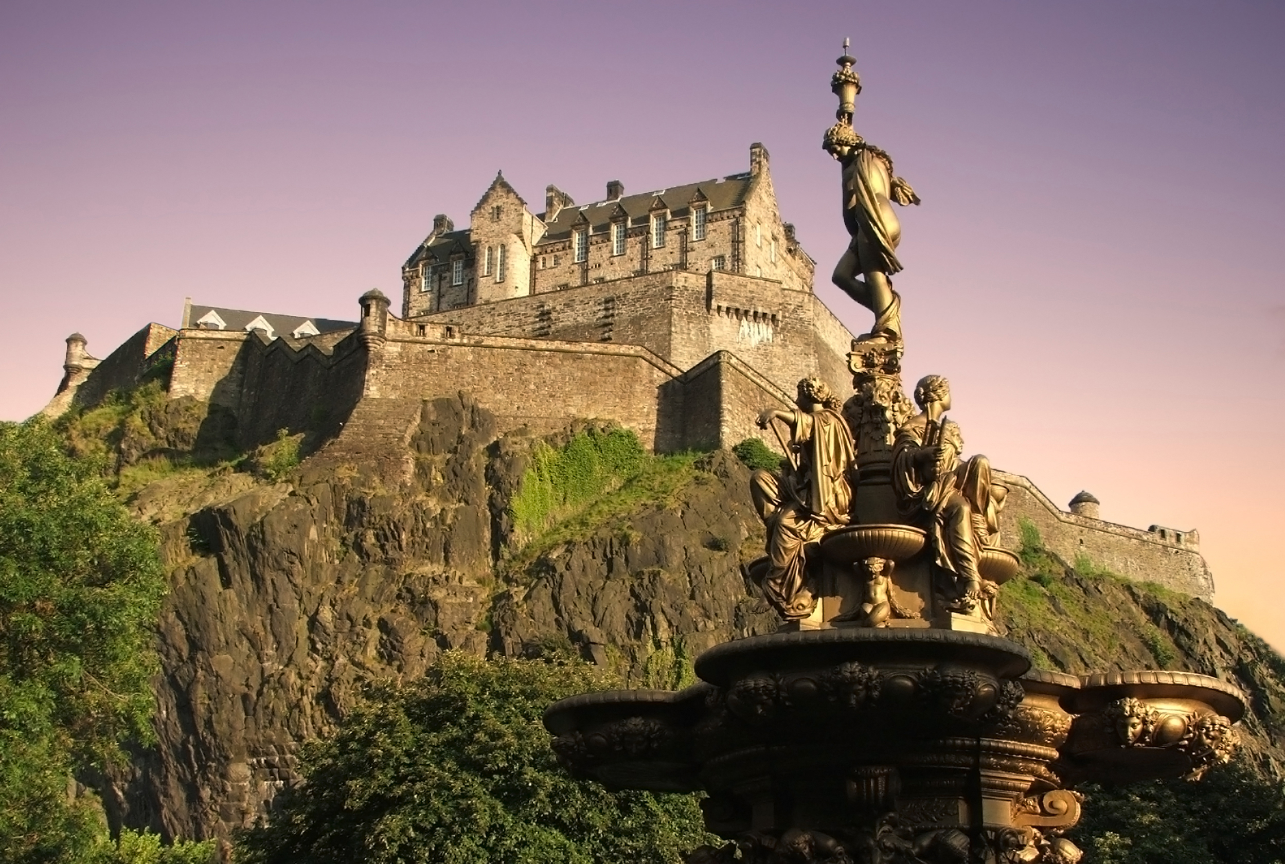 Det ikoniske Edinburgh Castle på vulkansk klippe ved tusmørke med forgyldt Ross-fontæne og frodig natur i Princes Street Gardens, Skotland