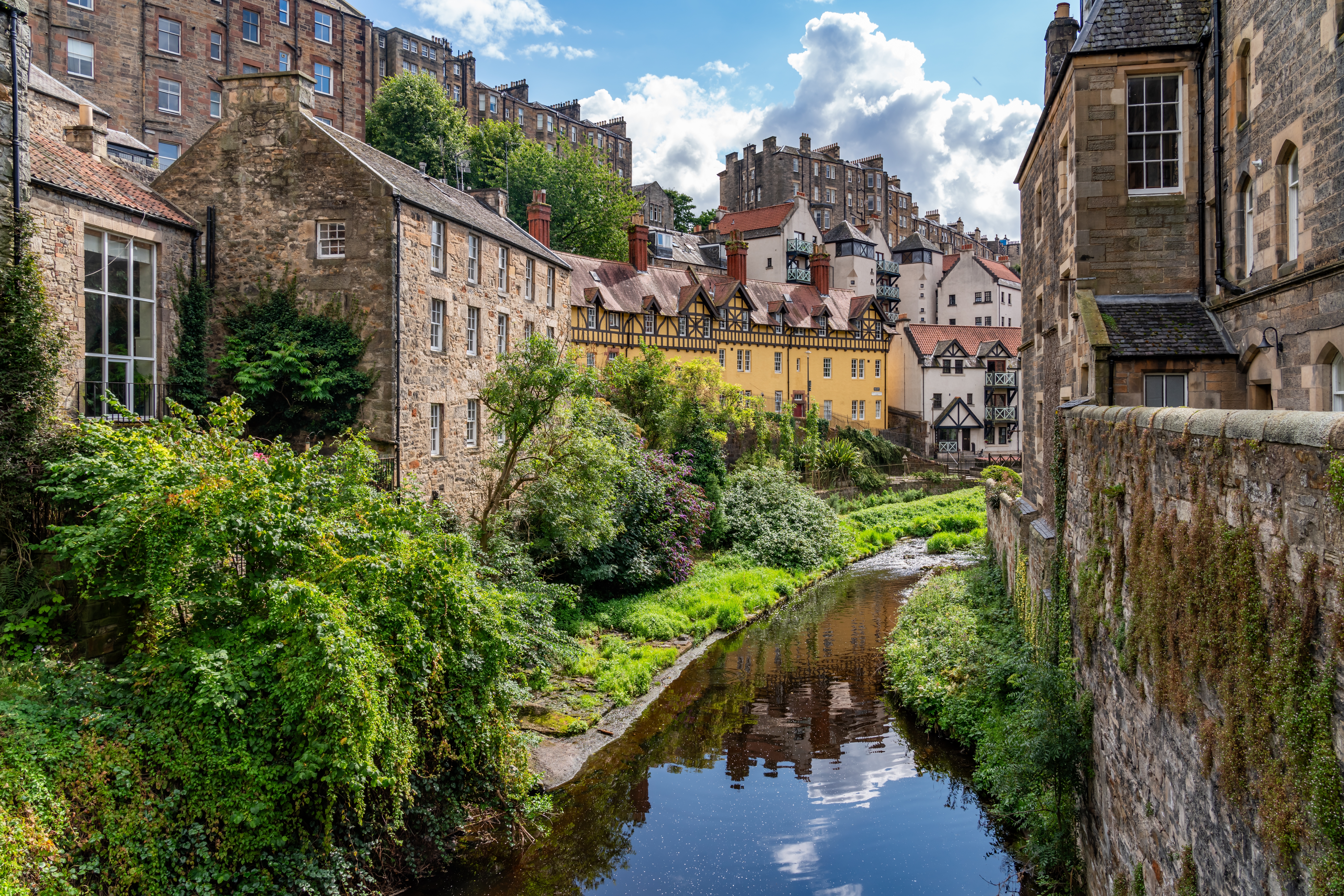 Charmerende udsigt over Dean Village i Edinburgh, Skotland, med historiske bygninger spejlet i den rolige Water of Leith omgivet af grøn vegetation