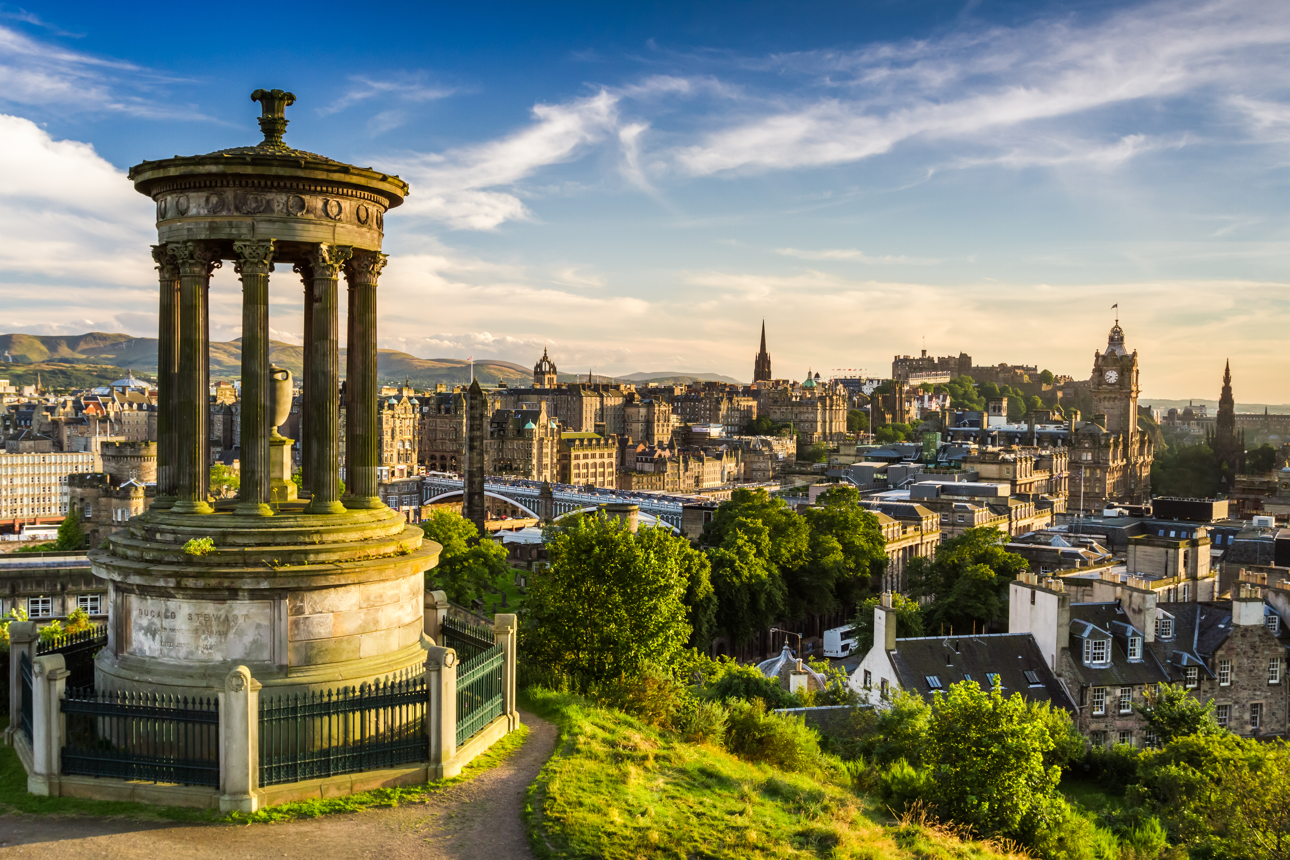 Storslået panorama over Edinburghs historiske skyline set fra Calton Hill med det klassiske Dugald Stewart-monument i forgrunden og det majestætiske Edinburgh Slot i baggrunden