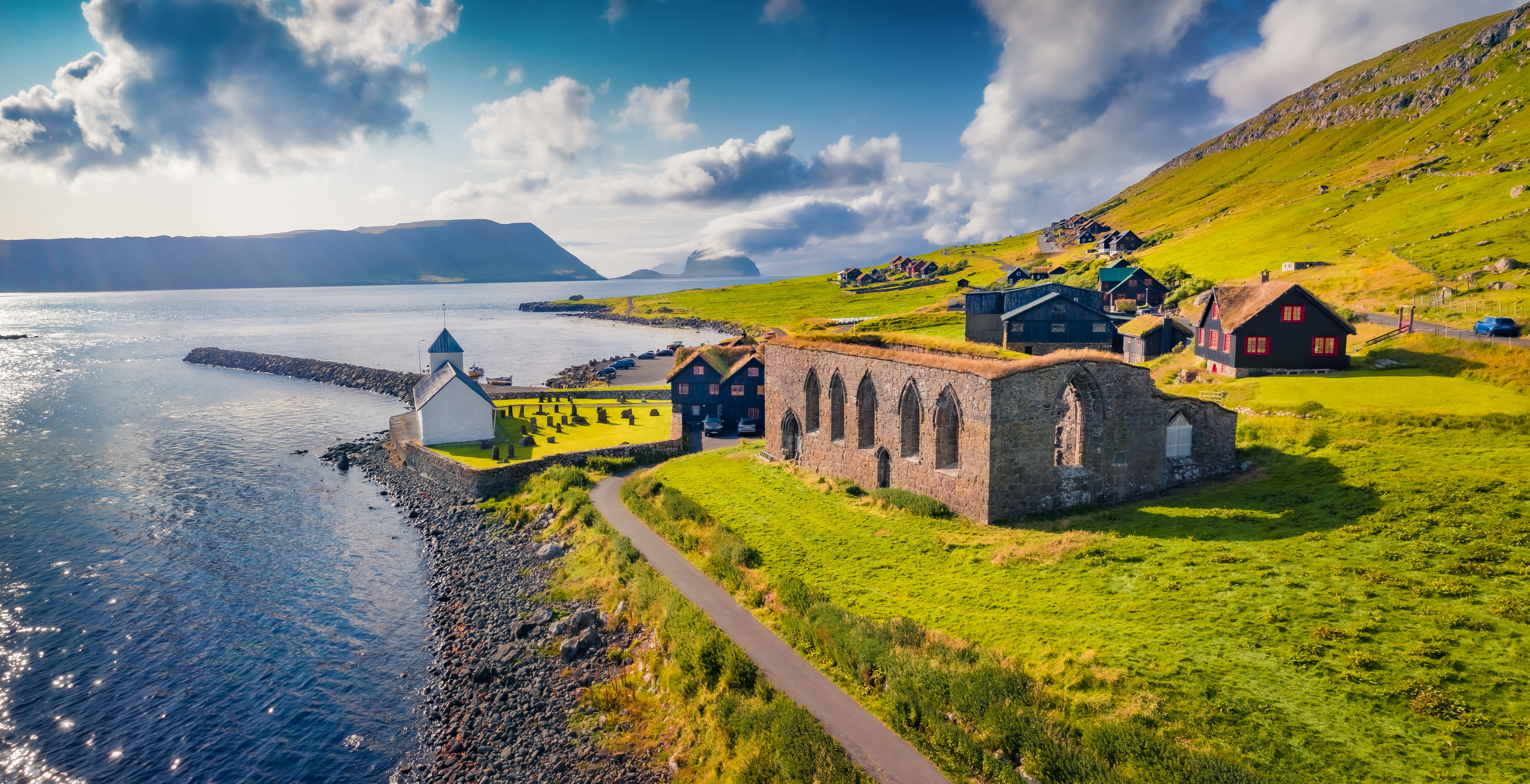 Fugleperspektiv over Kirkjubøur på Streymoy, Færøerne, med storslåede kirkeruiner, traditionelle nordatlantiske bygninger og smukt grønt bjerglandskab ved havet