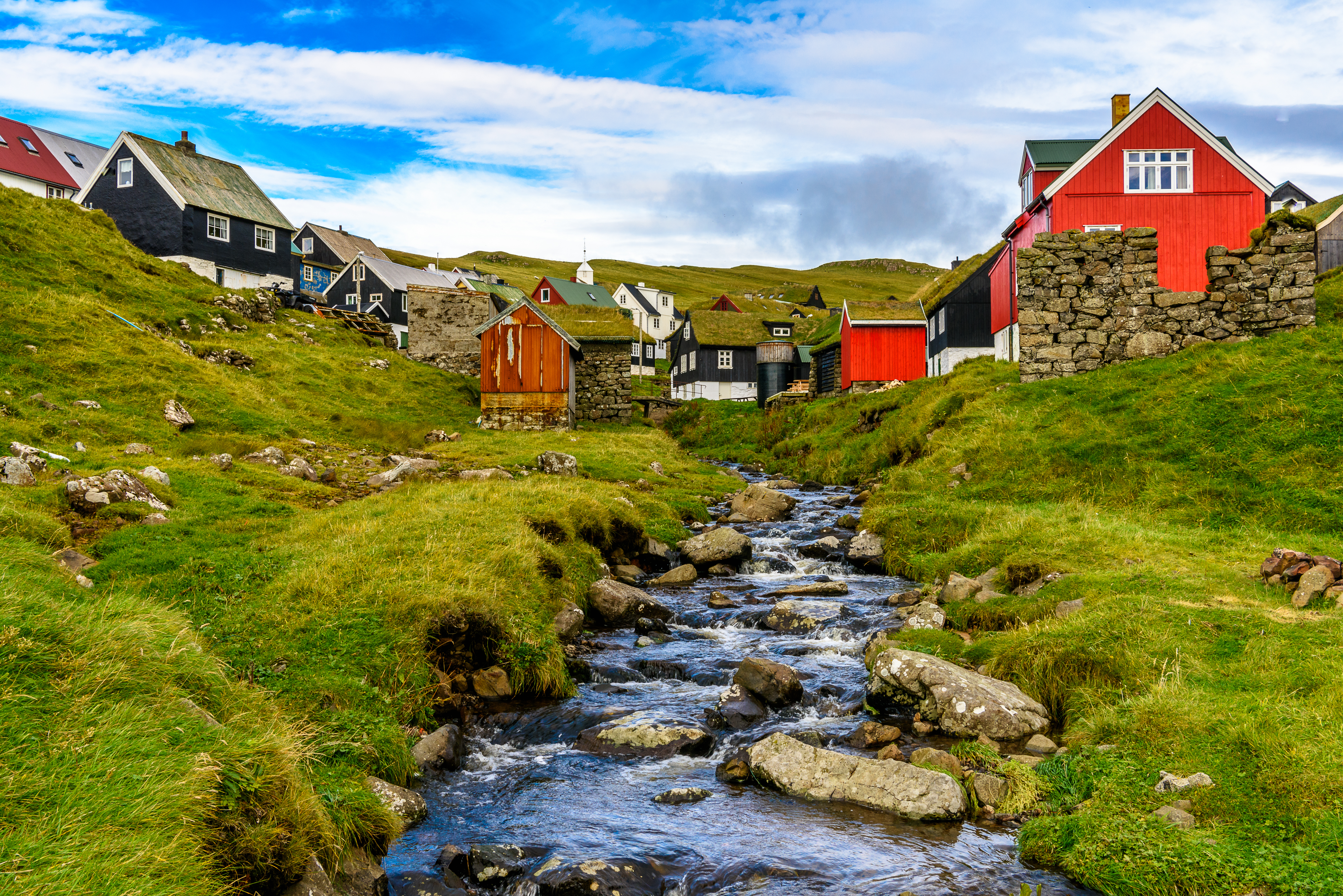 Stemningsfuld landsby på Færøerne med karakteristiske røde og sorte huse, natursten, bæk og bølgende grønne bakker