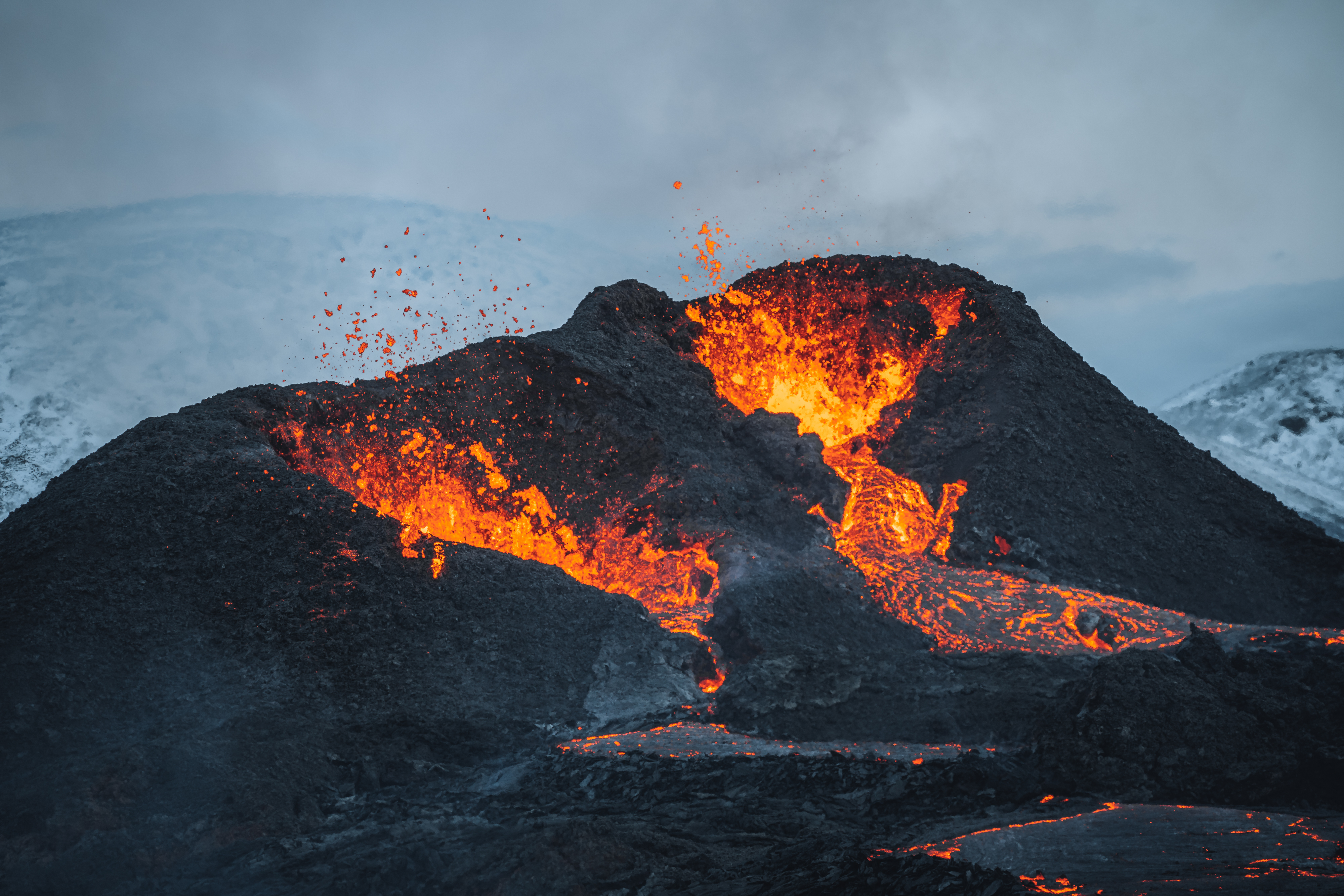 Naturfænomen på Island ved Fagradalsfjall vulkanen med kraftigt udbrud af glødende lava og magma fra krateret omgivet af sort vulkansk sten og snedækkede bjerge